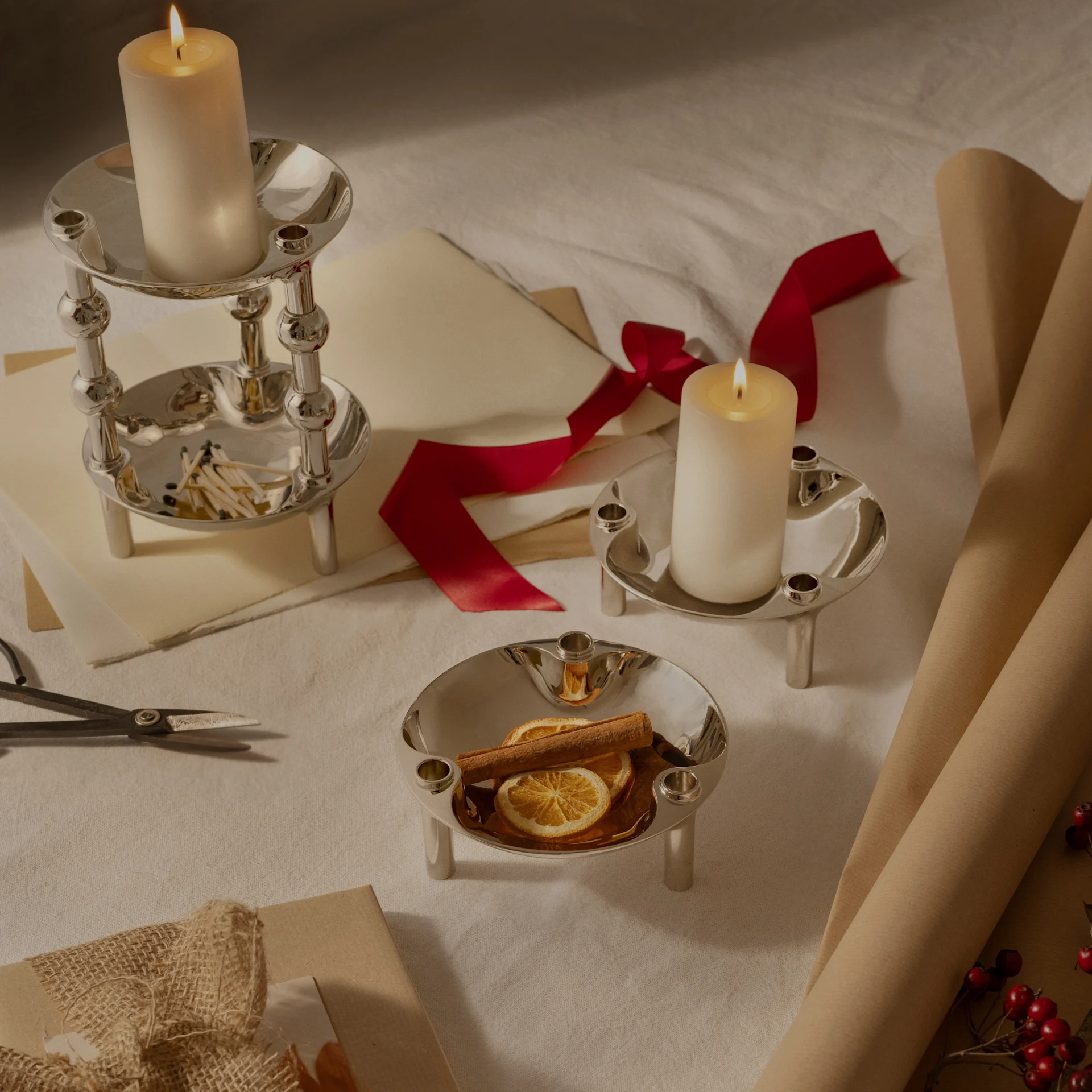 Three silver candle holders, two with lit pillar candles, one with dried oranges and cinnamon, arranged with wrapping paper and ribbon.