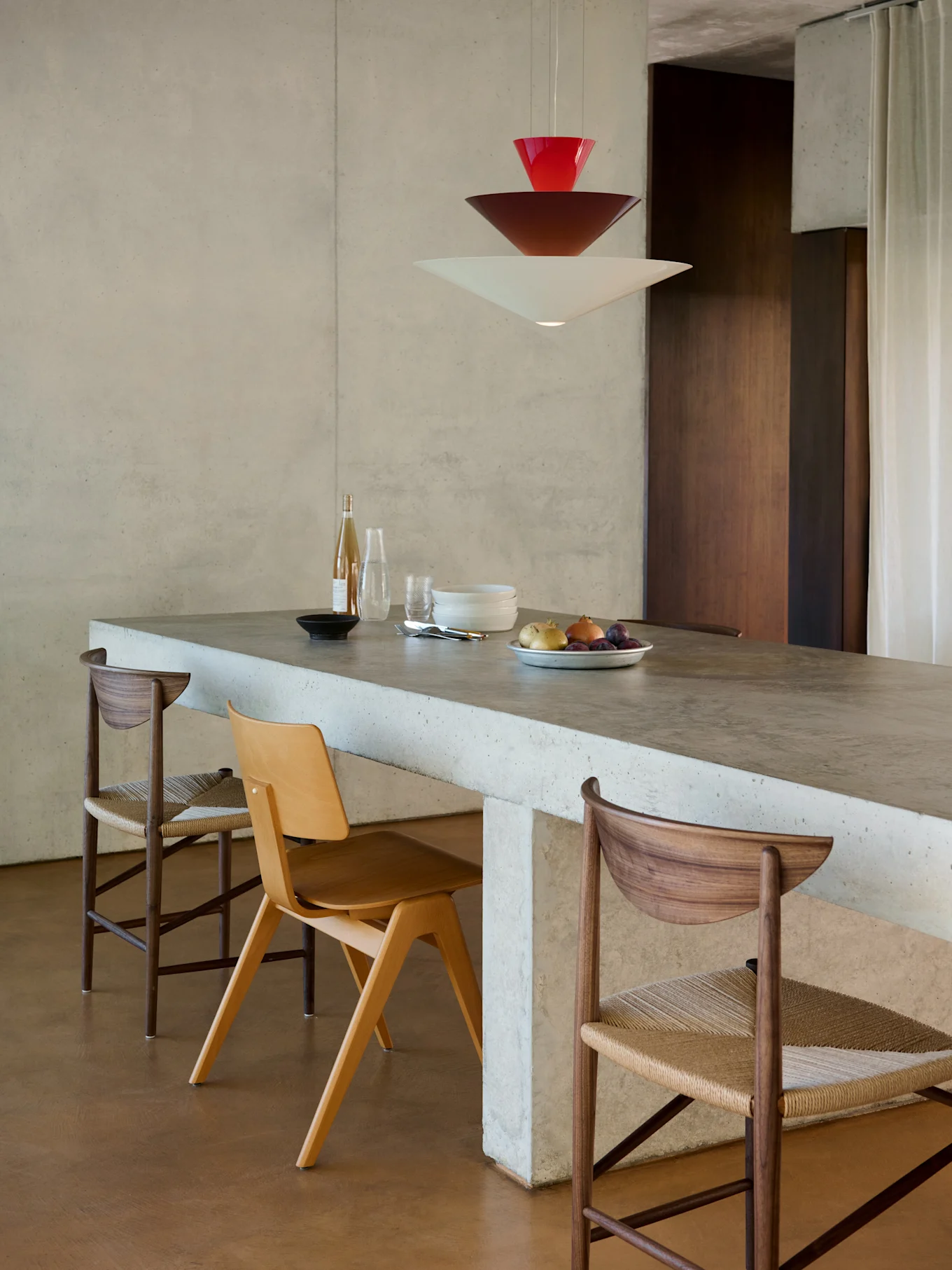 Modern dining area with concrete island, three chairs, and a stacked red, brown, white pendant light.