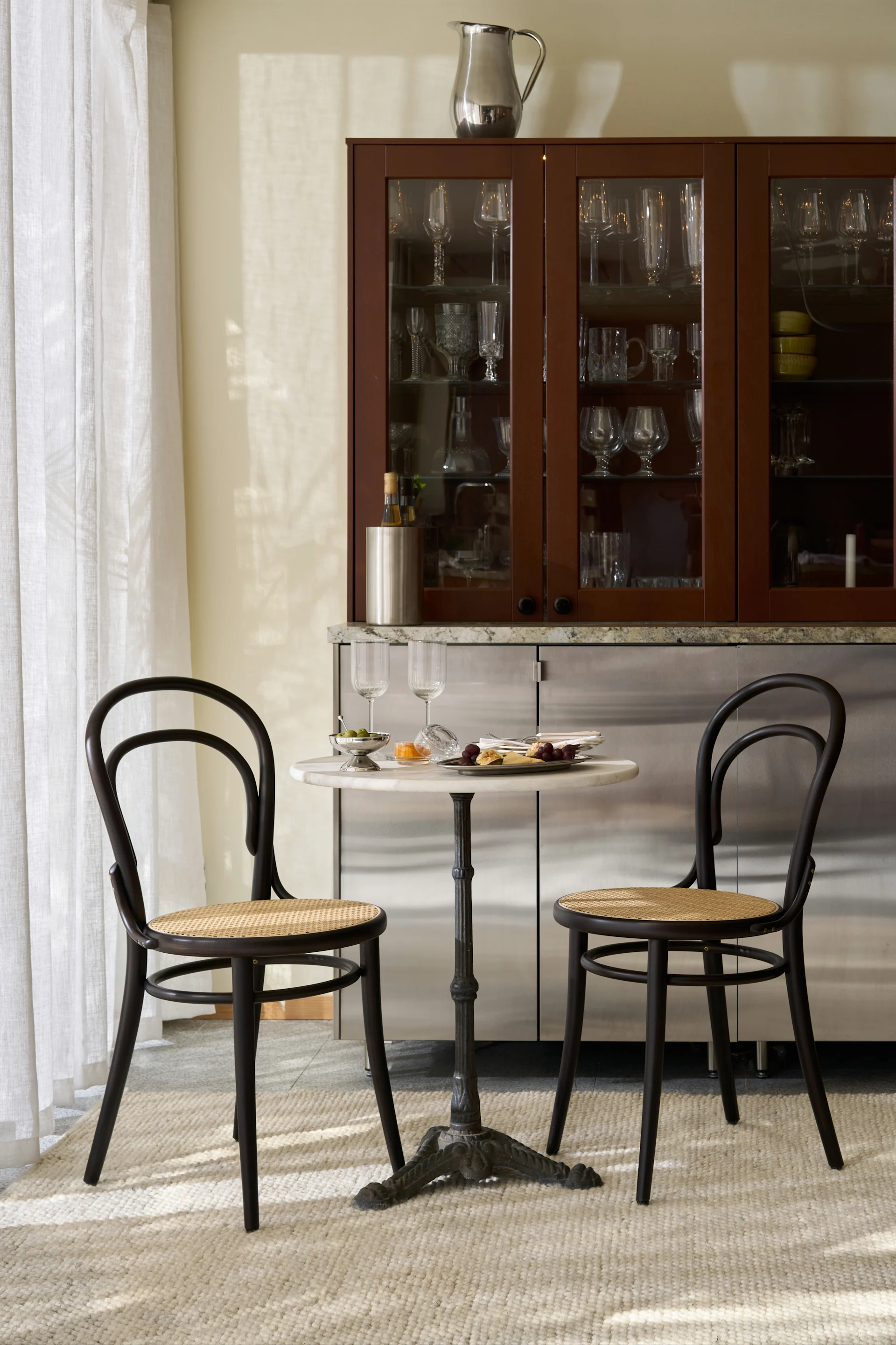 A stylish dining nook with a marble bistro table, two dark wood chairs, and a bar cabinet filled with glassware.