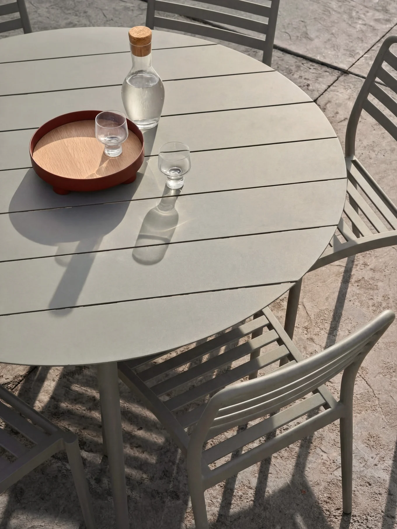 A light grey outdoor dining set with a round slatted table and chairs. On the table, a carafe of water and glasses on a tray.