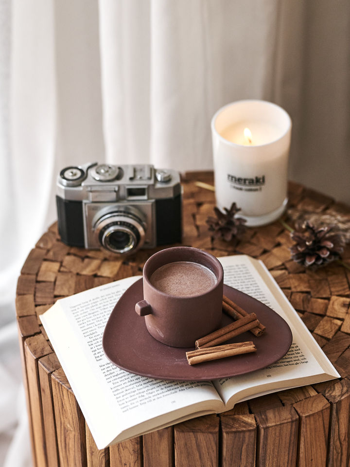 Brown Clay coffee cup from By On rests on top of a book on a side table in a cosy reading corner.