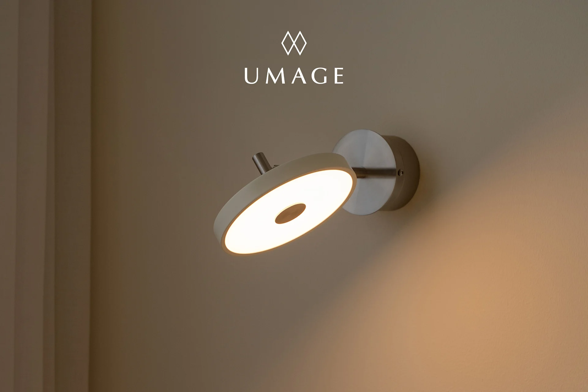 A modern circular wall lamp with a warm glowing light, mounted on a light brown wall. UMA logo above.