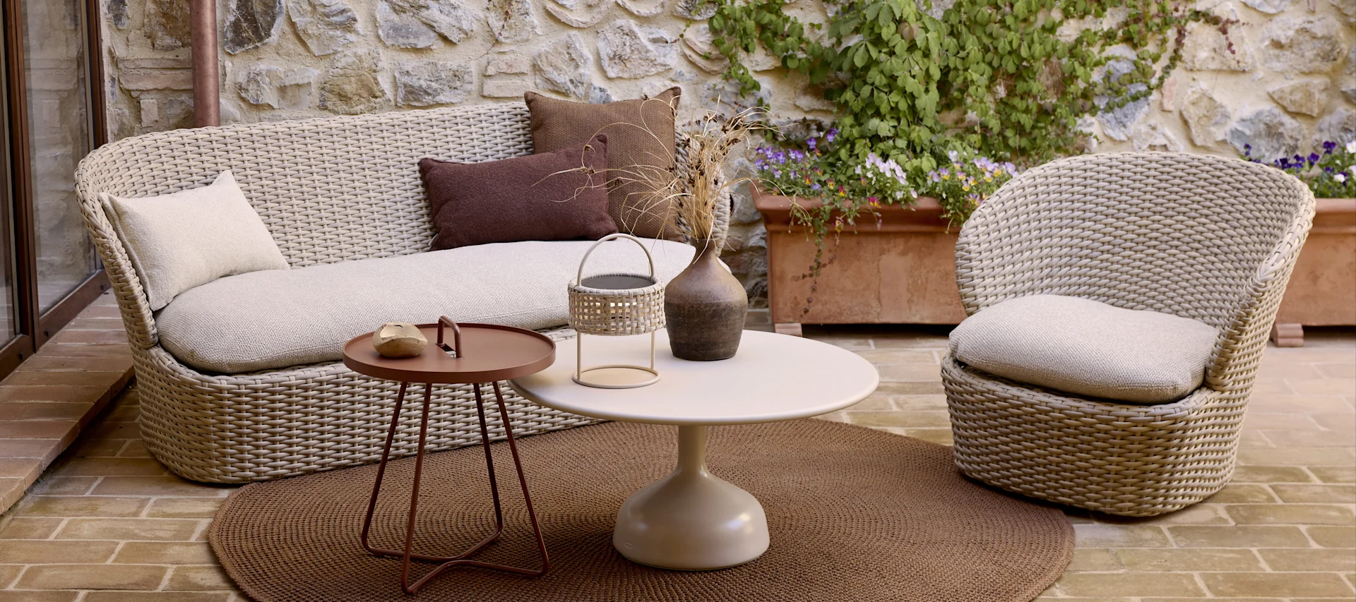Outdoor patio with light wicker sofa and armchair, two round tables, and a brown rug.
