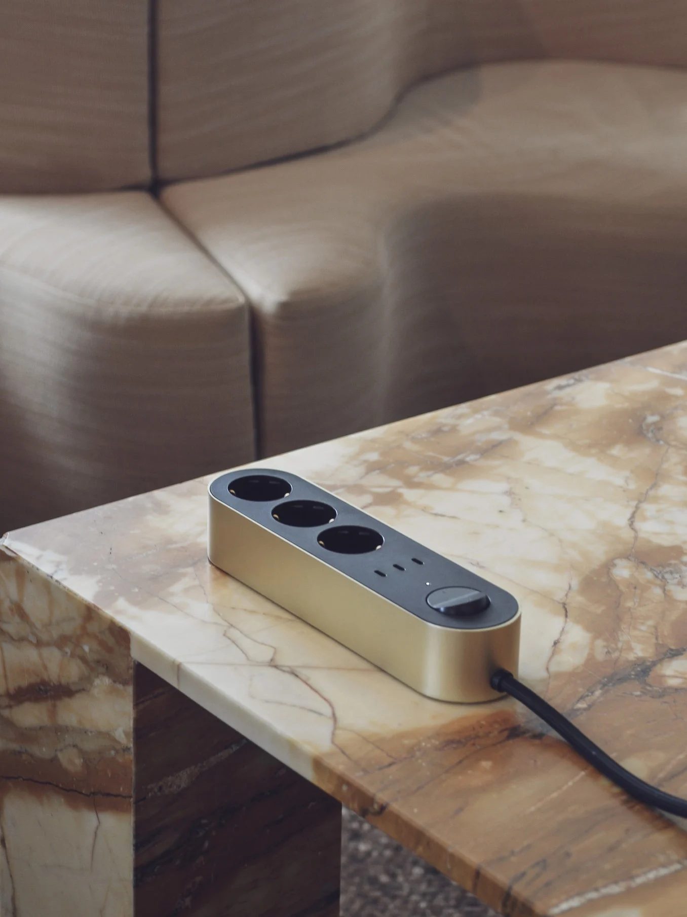 A gold power strip with three outlets and a switch sits on a marble table, with a curved sofa in the background.