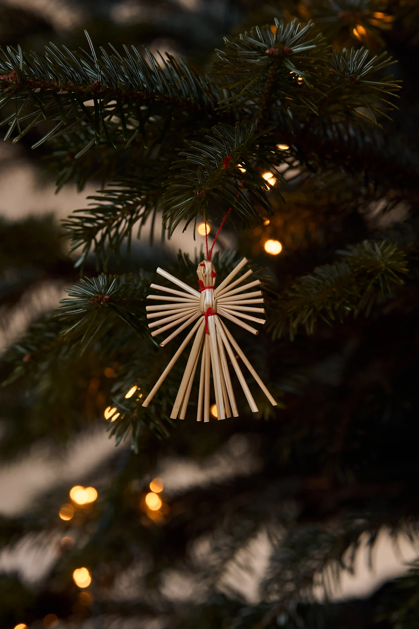 Straw angel ornament hanging on a Christmas tree branch with blurred lights in the background.