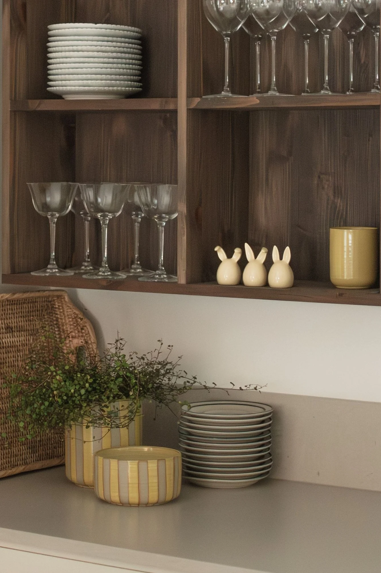 Wooden shelves with stacked white plates, various glasses, three ceramic bunnies, and a yellow mug. On the counter, a woven basket, plants in striped pots, and more plates.