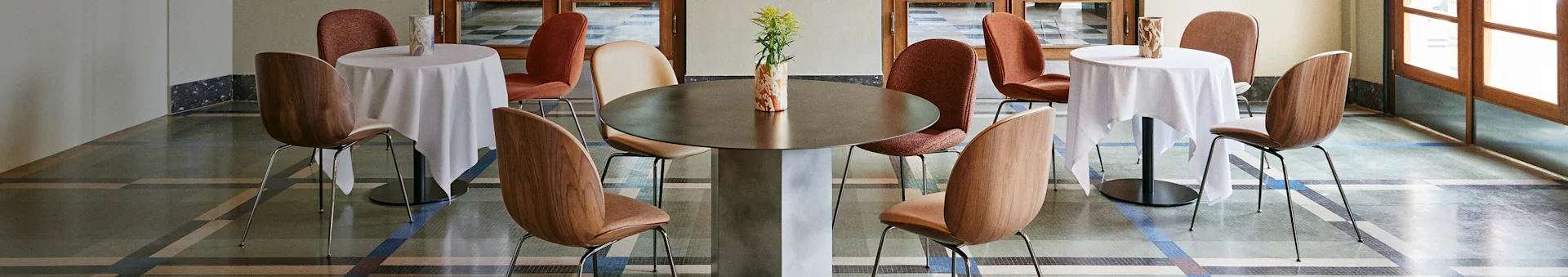 A stylish cafe interior with a large central table, two smaller tables with white cloths, and various chairs on a patterned floor.