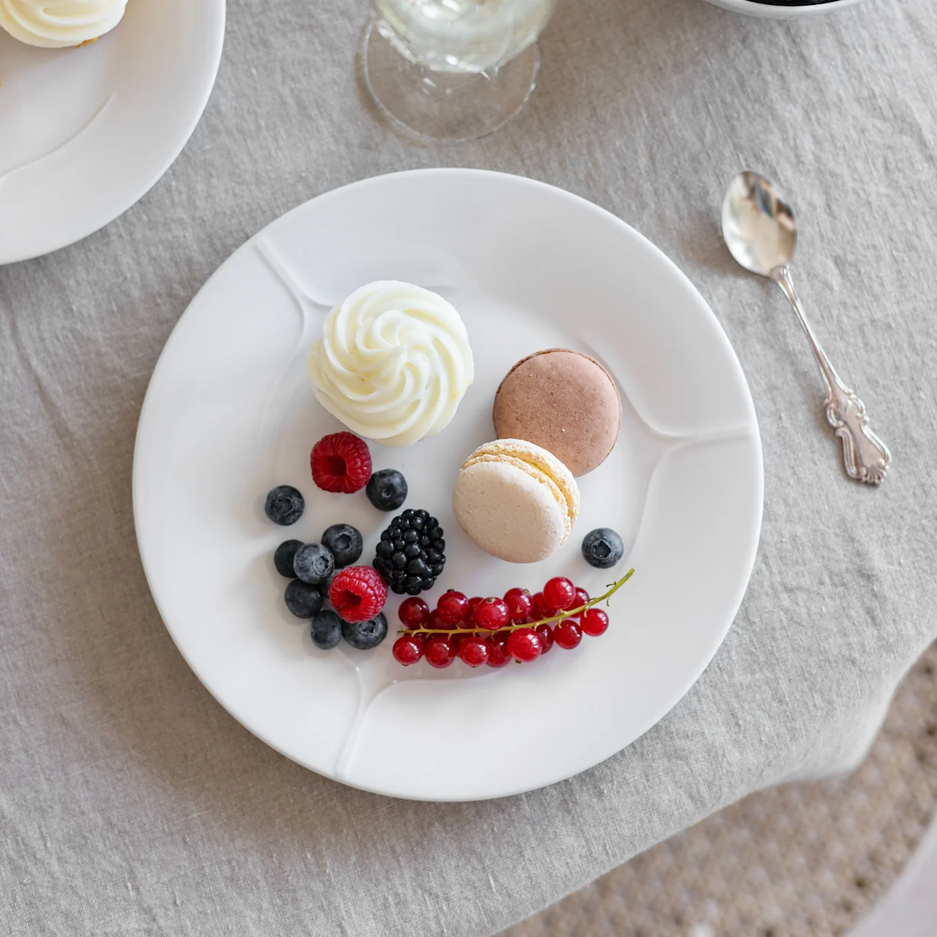 A white plate with a cupcake, two macarons, raspberries, blueberries, a blackberry, and red currants on a linen tablecloth.