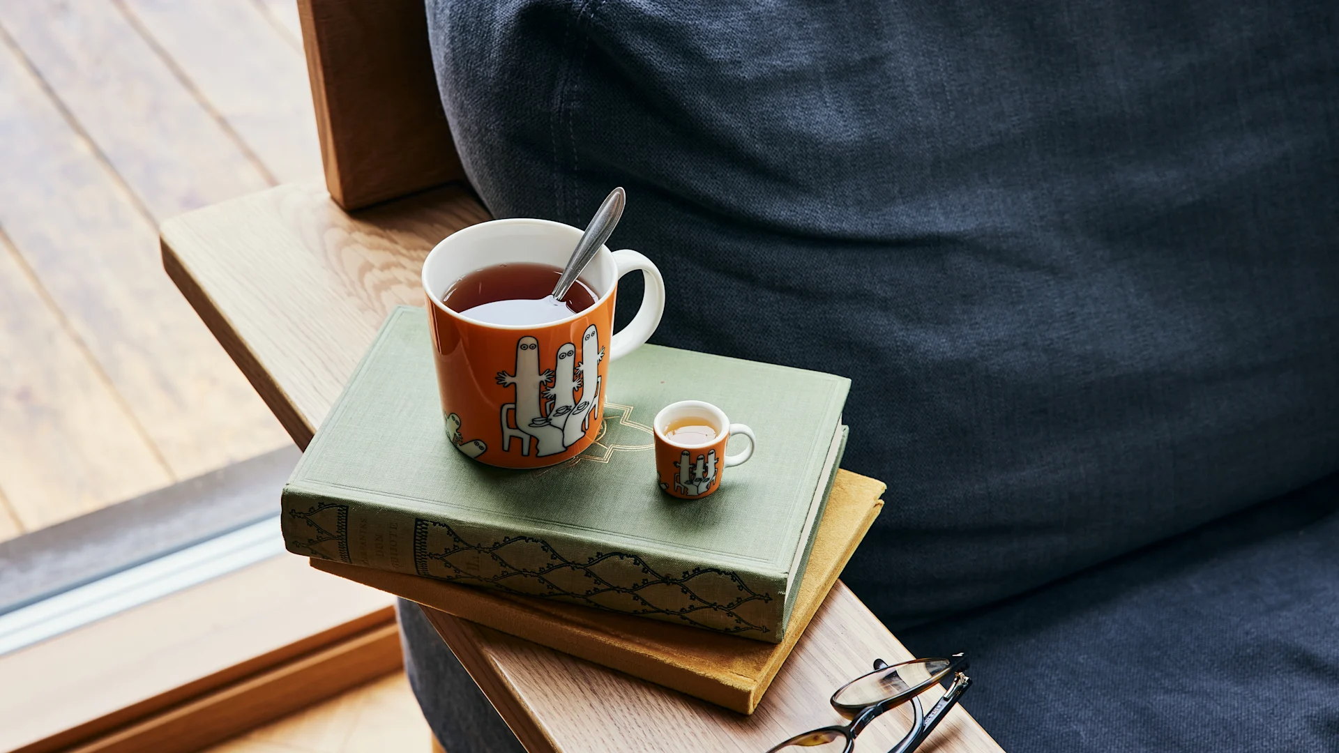 The orange-coloured Moomin character mug Hatifnatten with tea stands next to a sofa on a pile of books.