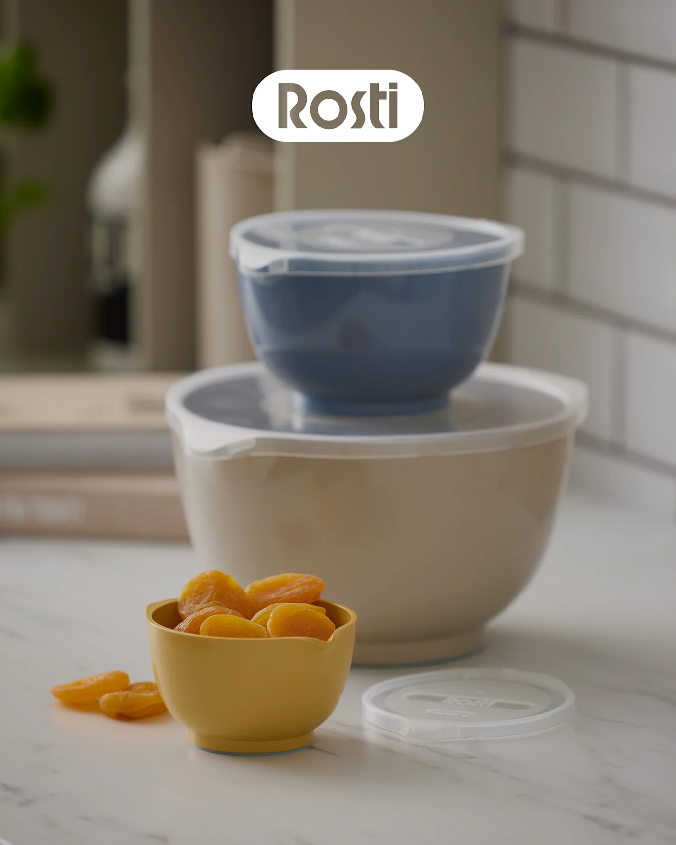 Three Rosti bowls on a counter: a beige bowl with a blue one stacked on top, both with lids, and a small yellow bowl filled with dried apricots.