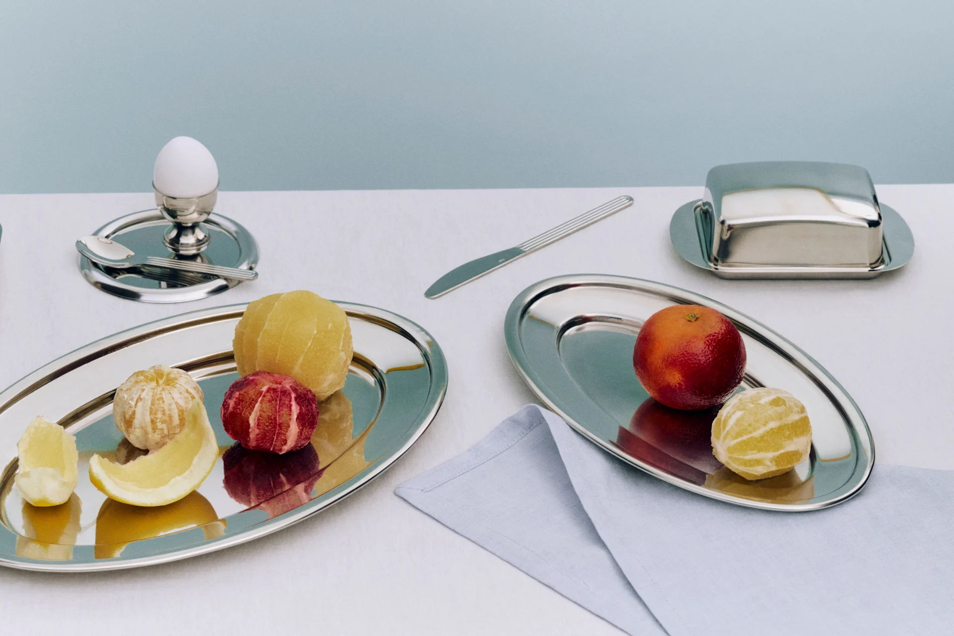 A breakfast setting with two silver trays holding peeled citrus fruits, an egg in an egg cup, a butter dish, and a knife.