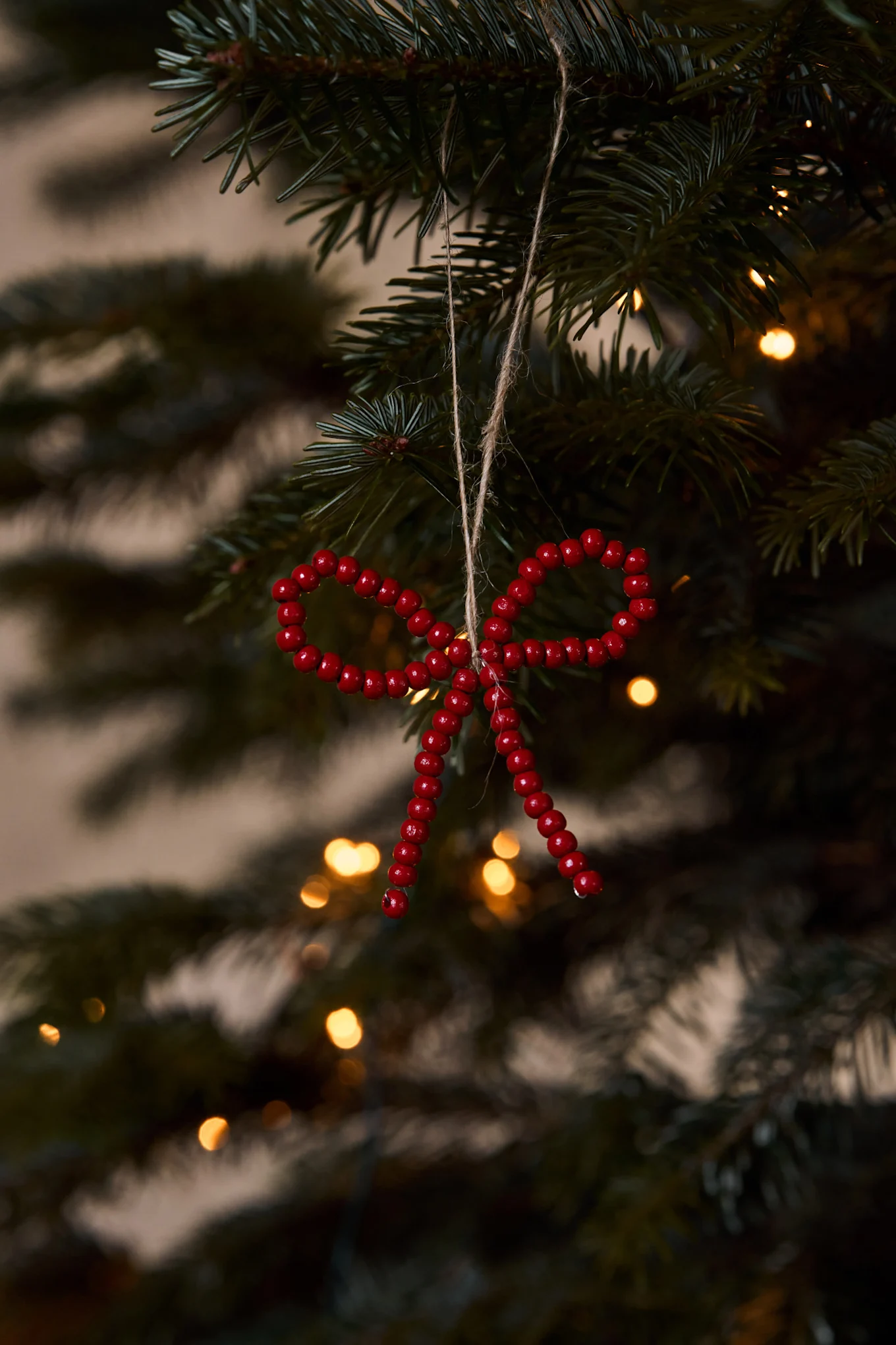 Close-up of a red beaded bow ornament hanging on a Christmas tree with blurred yellow lights.