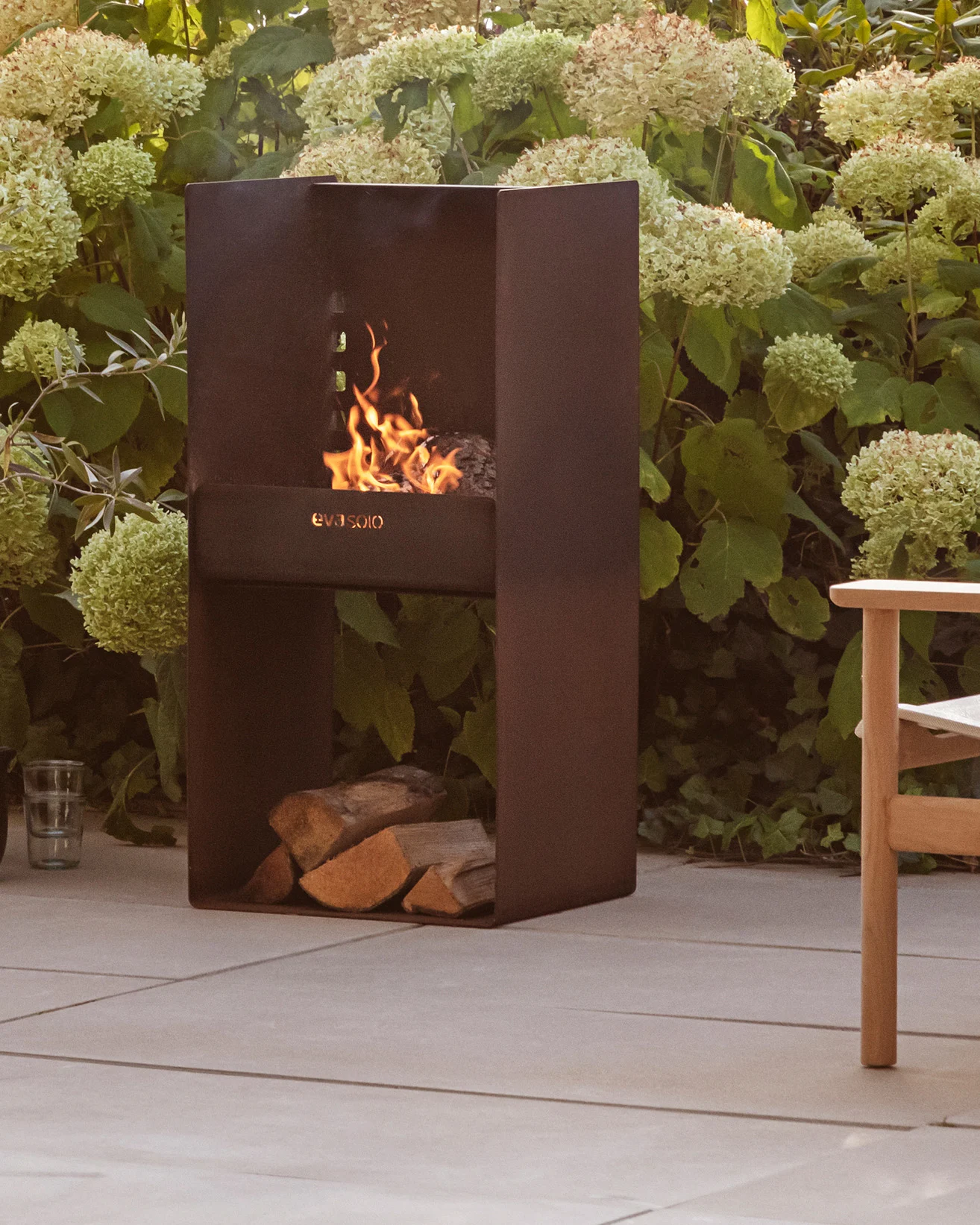 Modern dark metal outdoor fireplace with a fire burning, stacked logs below, surrounded by green plants and a patio.