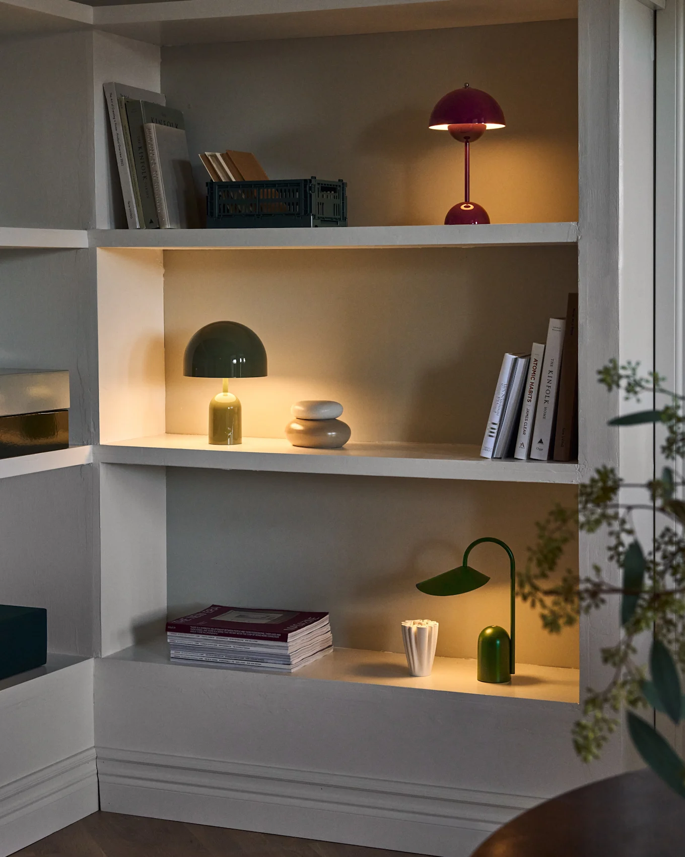 Three illuminated shelves in a built-in unit, displaying red and green mushroom lamps, a green desk lamp, books, and decor.