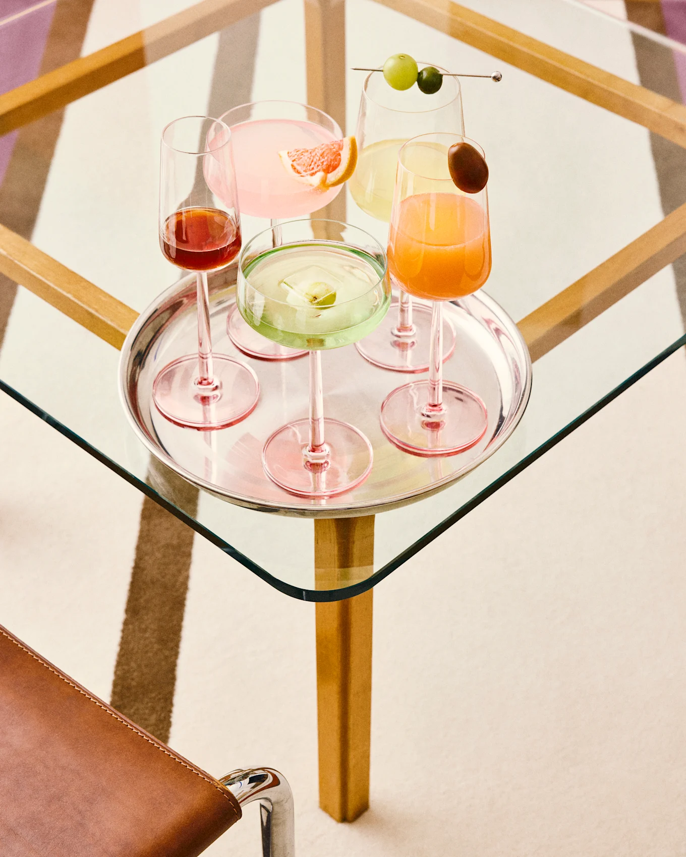 Five colorful cocktails on a silver tray: pink with grapefruit, green with fruit ice, yellow with grapes, orange with a garnish, and dark red, all on a glass table.