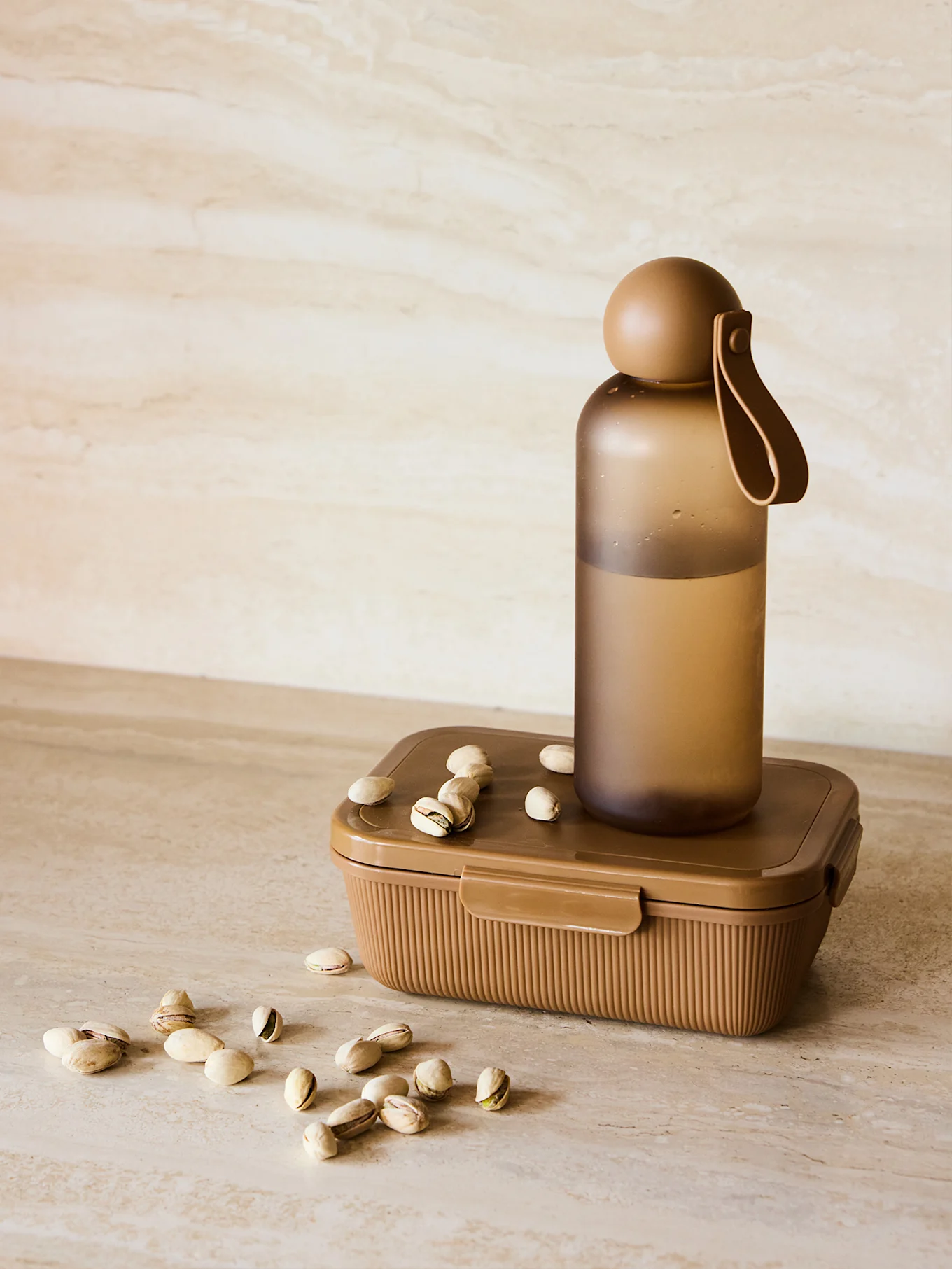 Brown water bottle on a ribbed lunch box with scattered pistachios on a marbled surface.