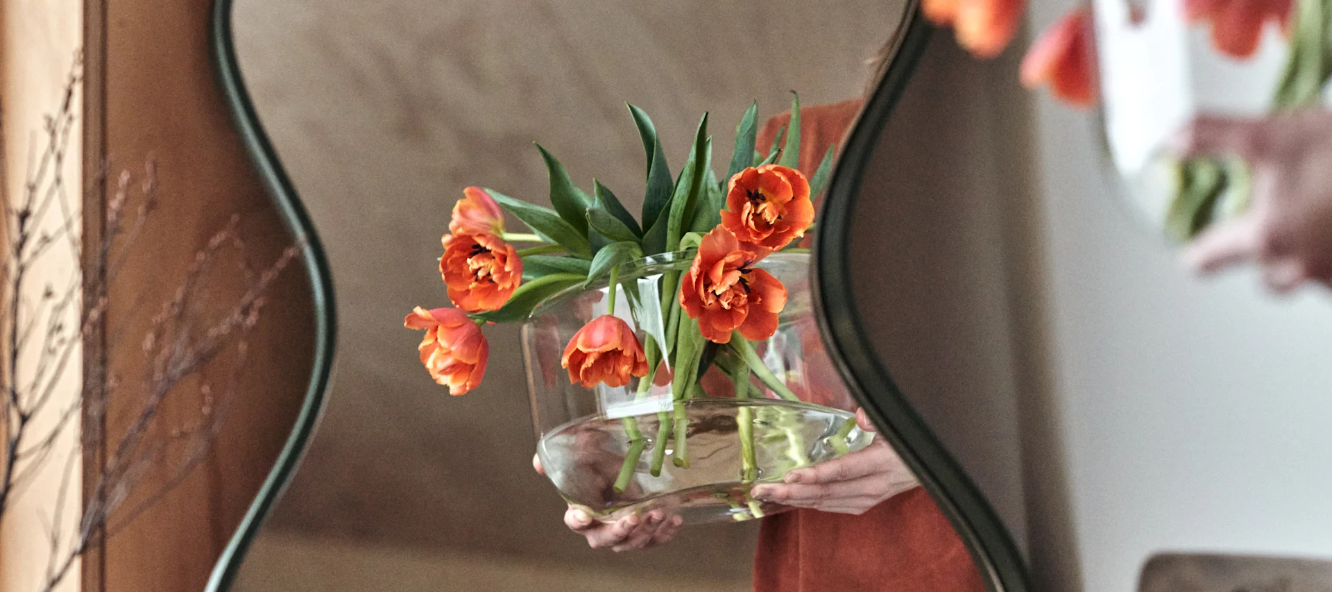A pair of hands carries a vase of fresh flowers arranged in a clear glass vase reflected in a curved mirror from Ekbacken Studios.