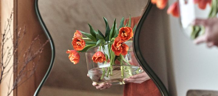 A pair of hands carries a vase of fresh flowers arranged in a clear glass vase reflected in a curved mirror from Ekbacken Studios. 