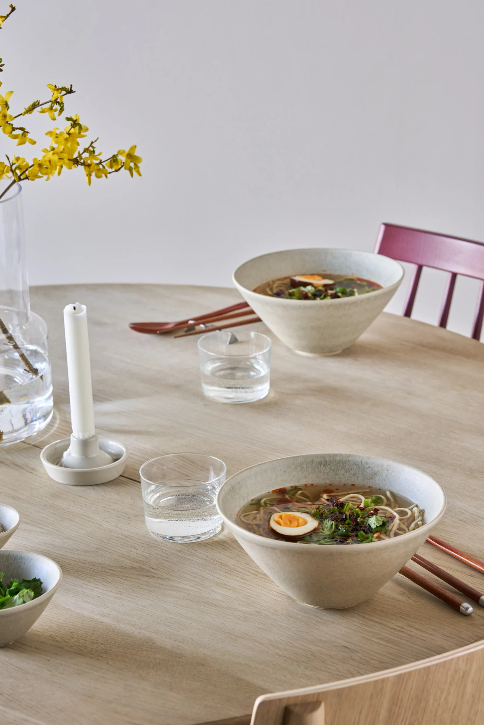 Two bowls of ramen with eggs and garnish on a light wooden table, with water glasses and yellow flowers in a vase.