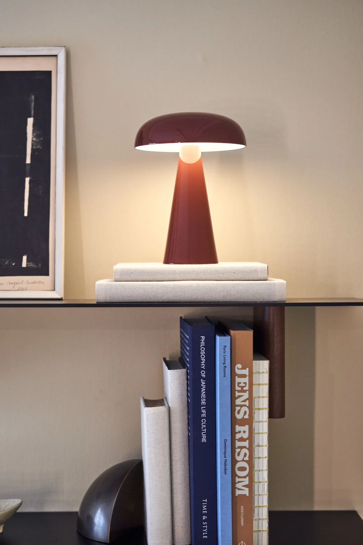 The cordless table lamp Como from &tradition sits on a shelf with books and a picture frame. 