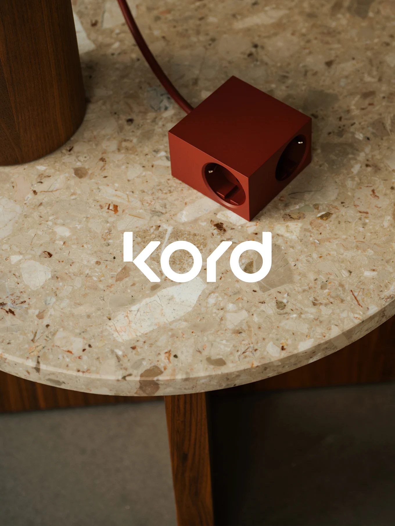 A dark red, cube-shaped power strip with two visible outlets rests on a round marble table, with the white "kord" logo.