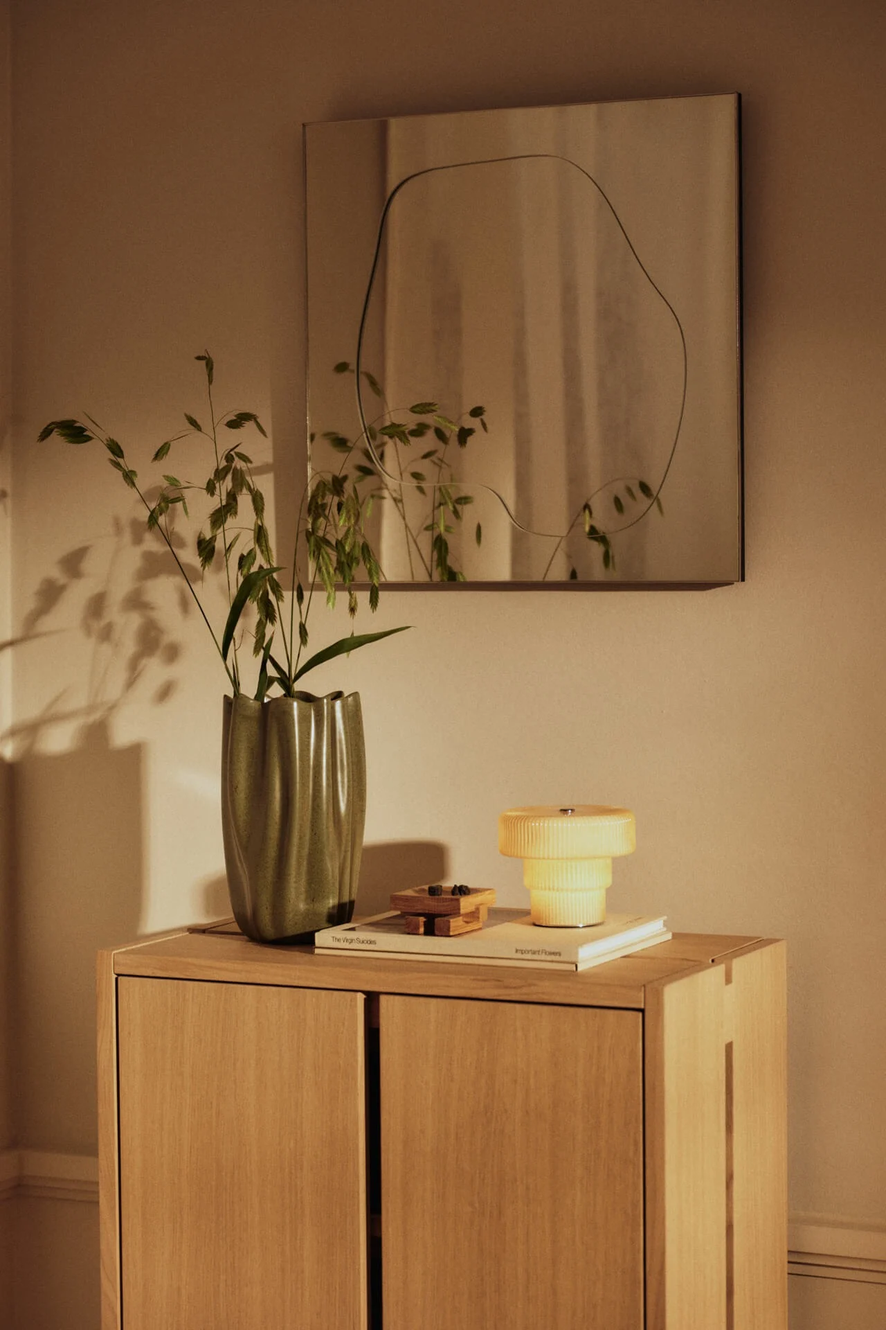A wooden cabinet with a green wavy vase of grass, a yellow tiered lamp, and a mirror with an irregular outline on the wall.