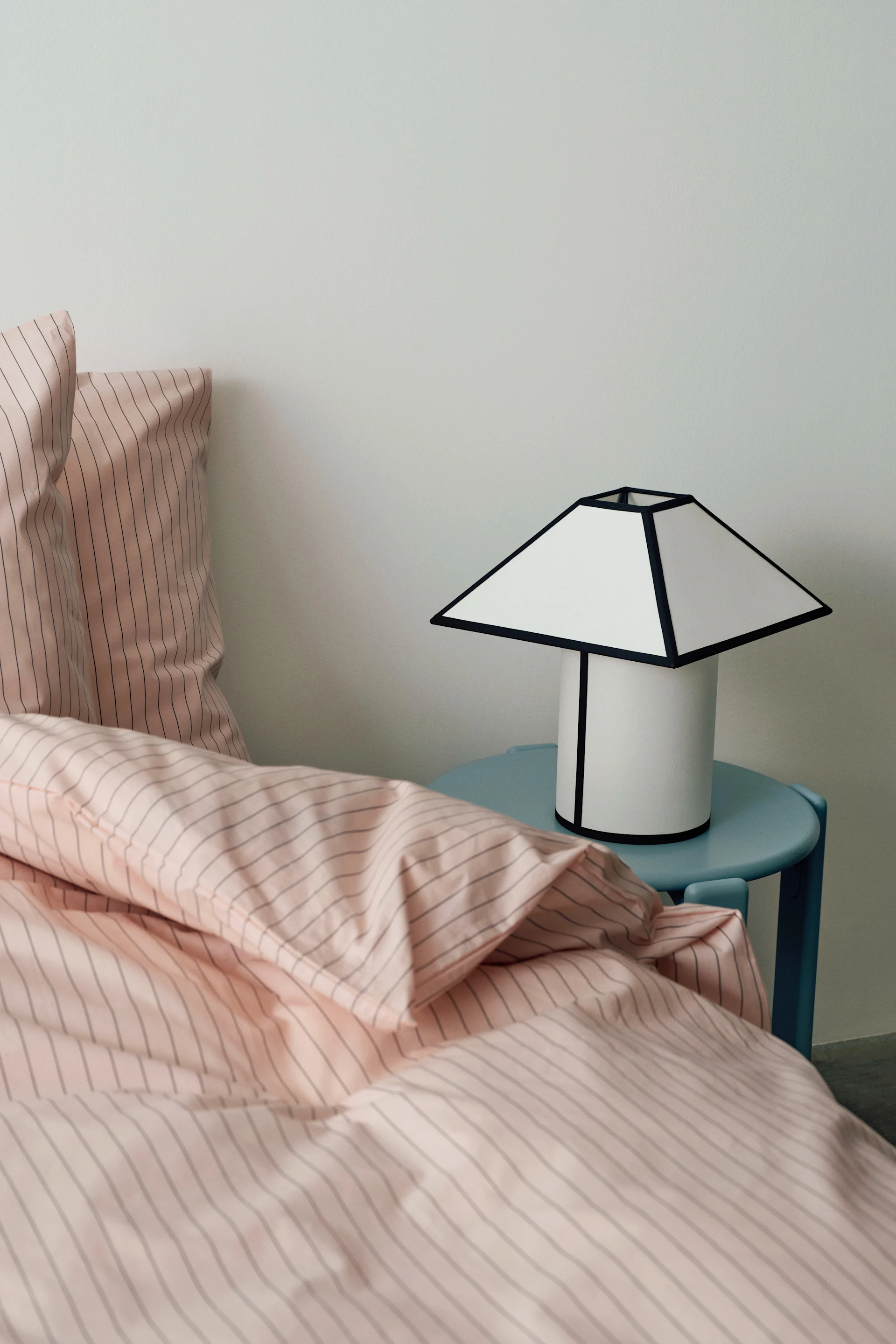 Pink striped bedding on a bed with a white lamp with a black-outlined shade on a light blue side table.