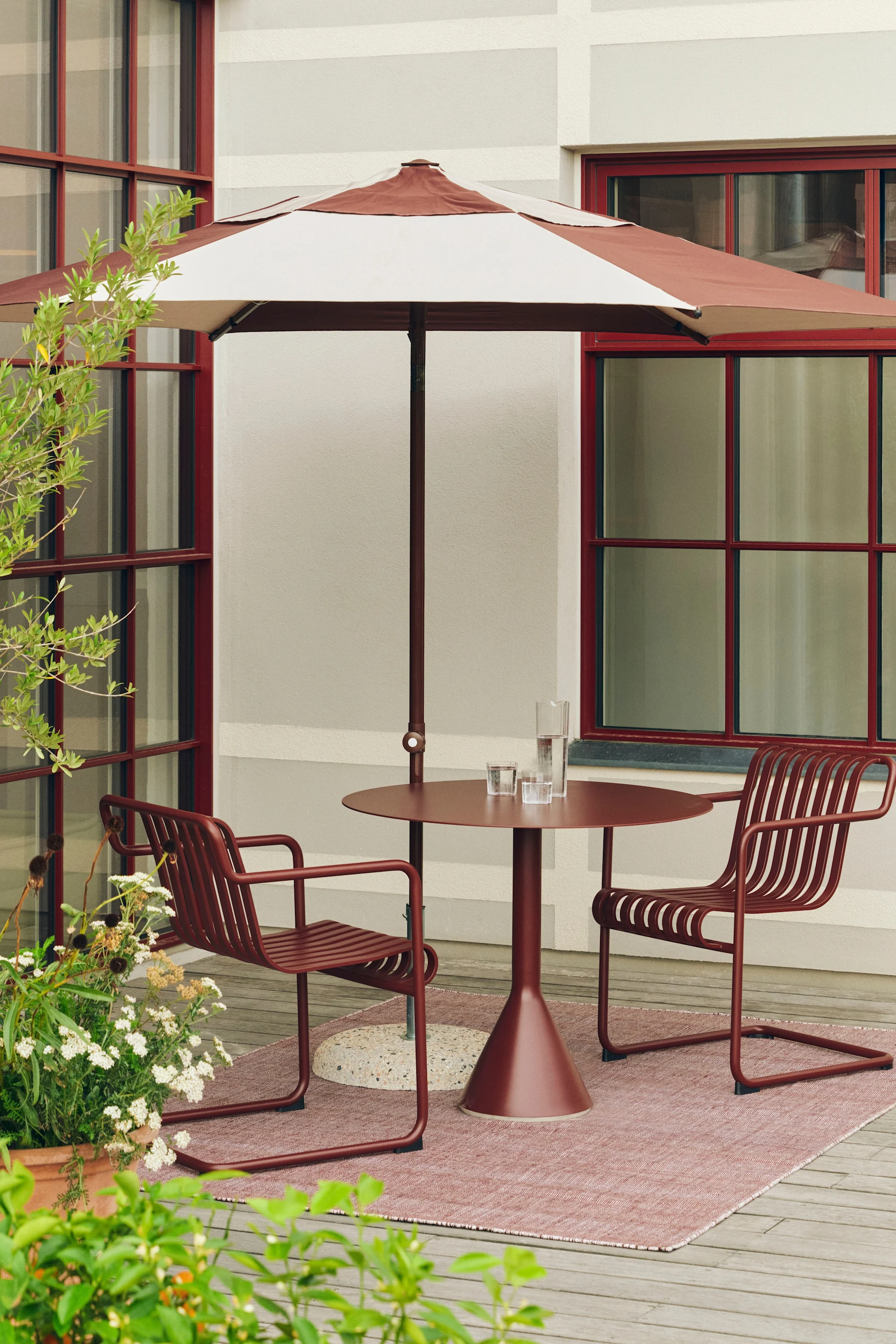 Outdoor patio with a red-brown striped umbrella, matching round table, and two slatted chairs on a pink rug.
