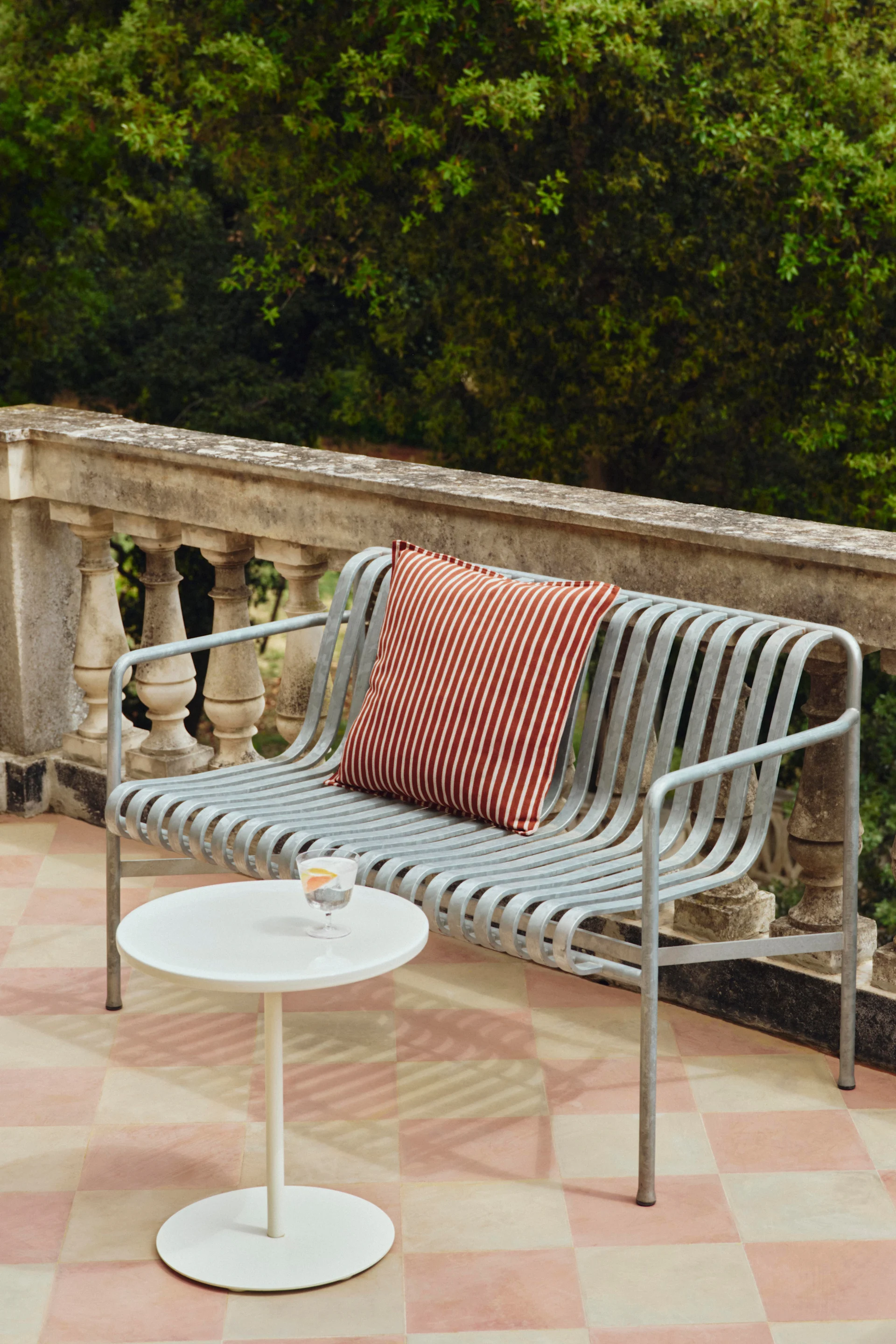 A light grey metal slatted outdoor bench with a red and white striped cushion, next to a white side table with a drink on a checkered patio.