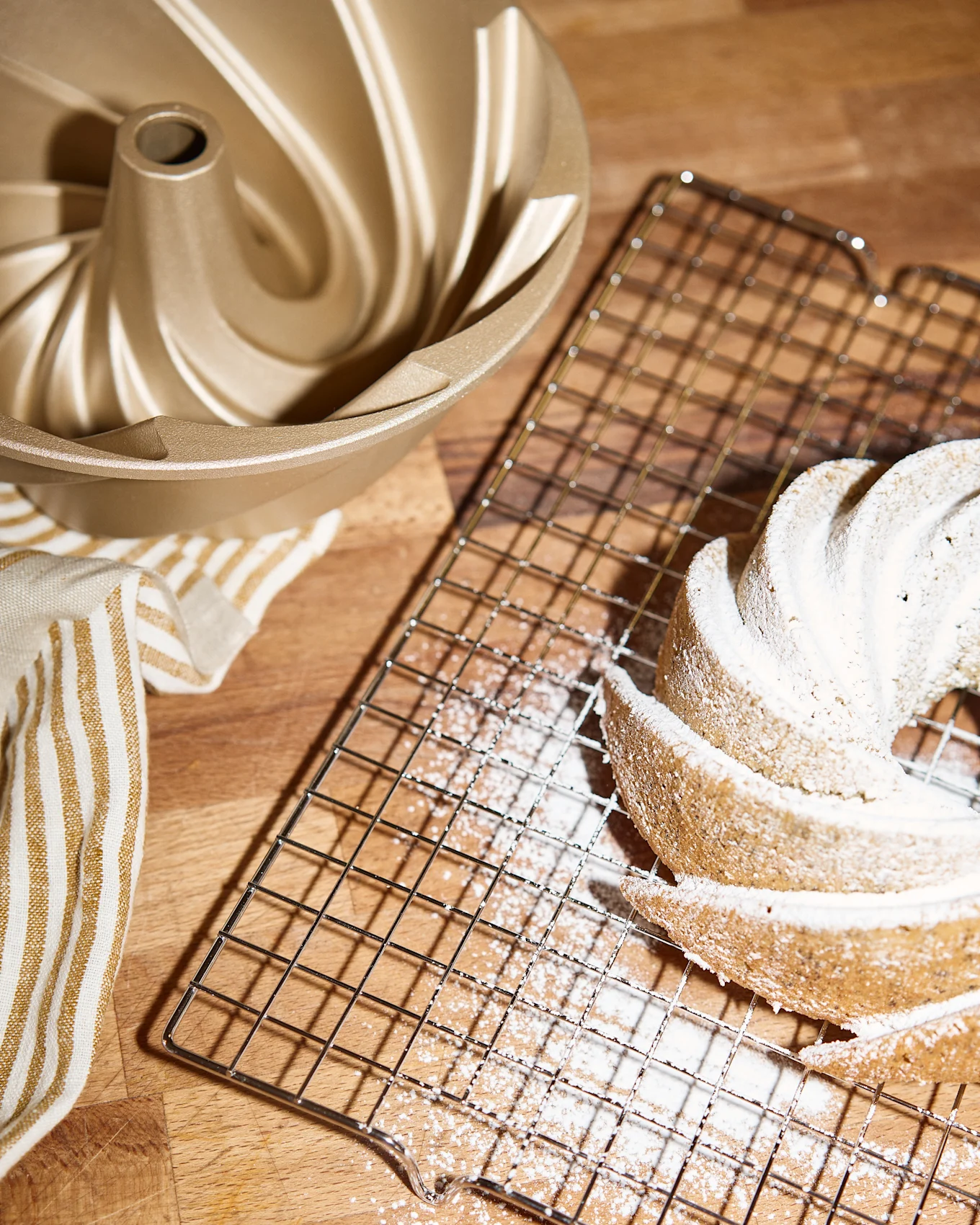 A golden bundt cake mold, a cooling rack with a powdered sugar-dusted bundt cake, and a striped towel on a wooden surface.