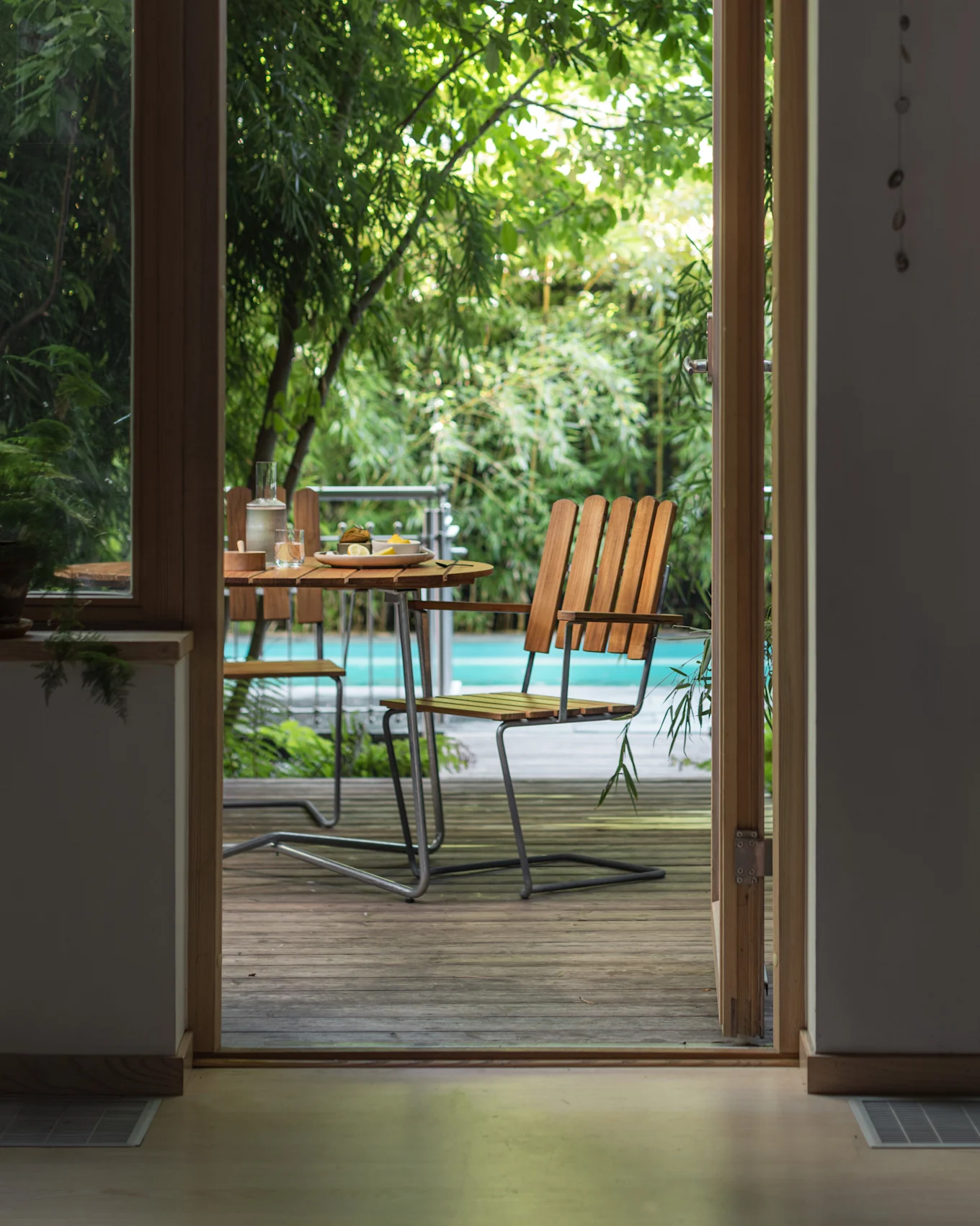 View through an open doorway to an outdoor patio with a wooden table, chairs, and a pool surrounded by green plants.