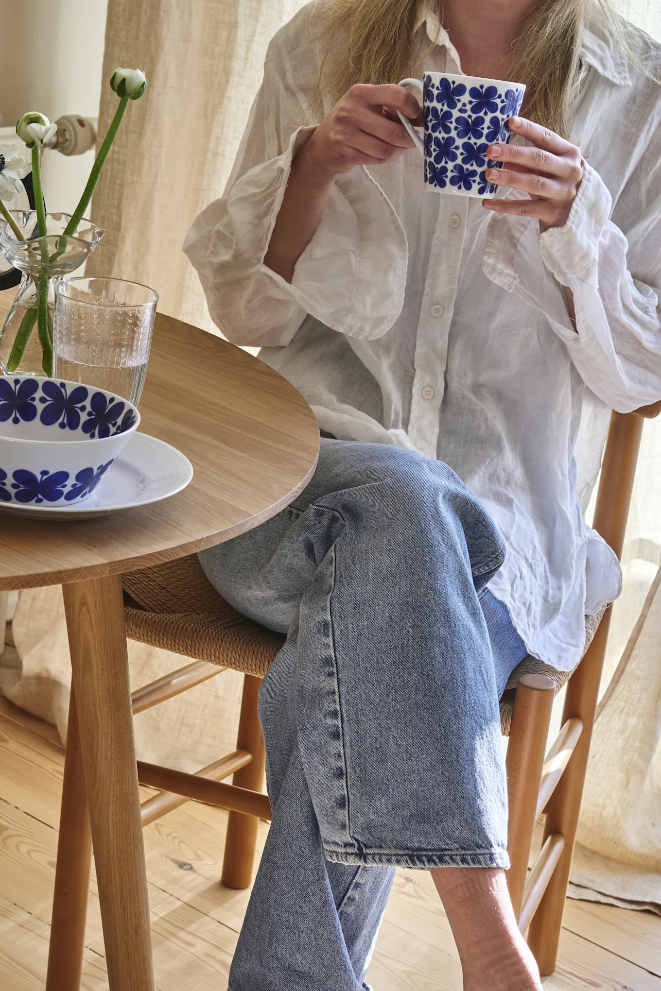 Person in white shirt and jeans holding a blue floral mug at a wooden table with a matching bowl and flowers.