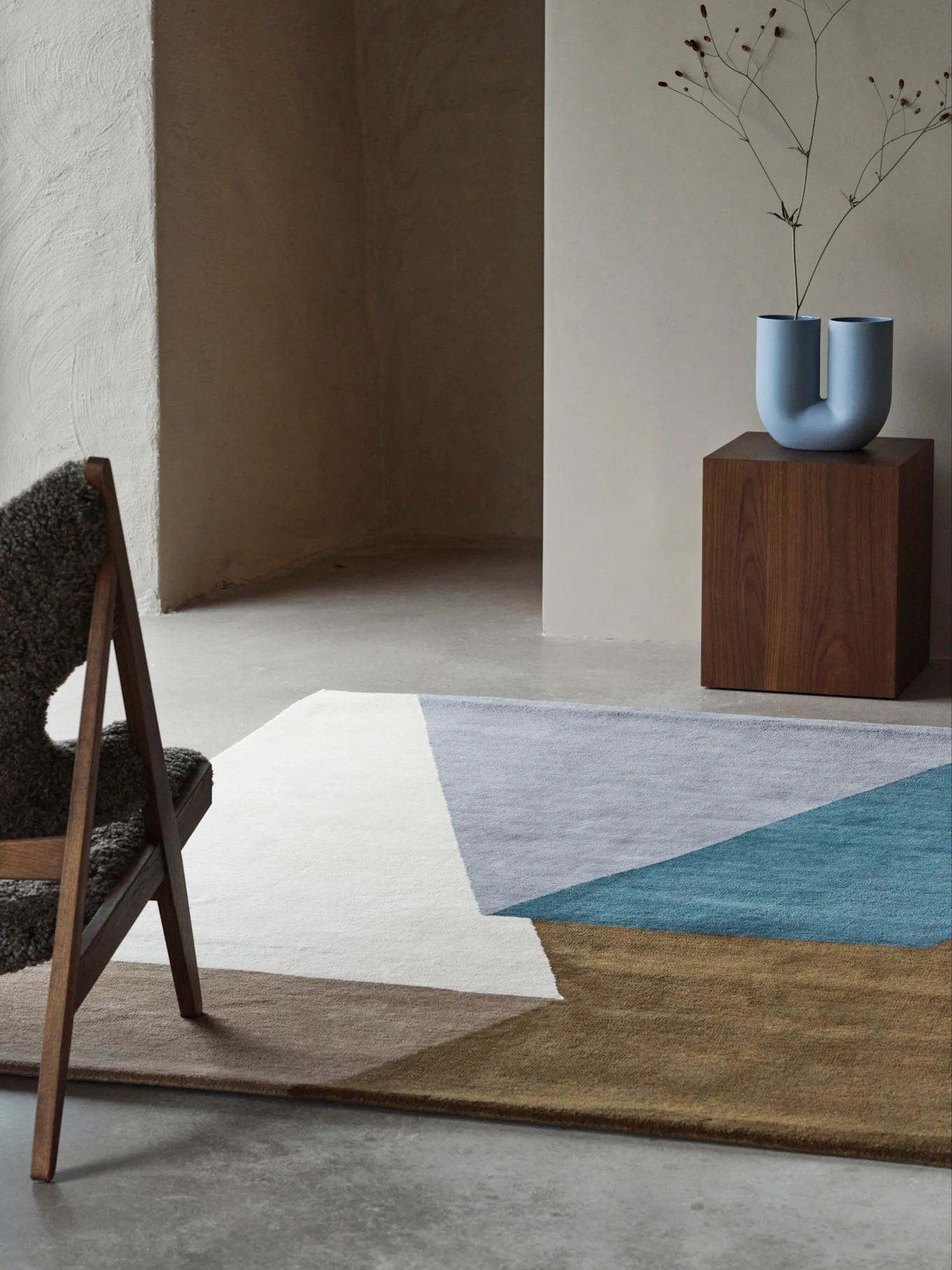 Modern room with a geometric rug in white, grey, teal, and brown, a U-shaped blue vase on a wooden pedestal, and a fluffy chair.