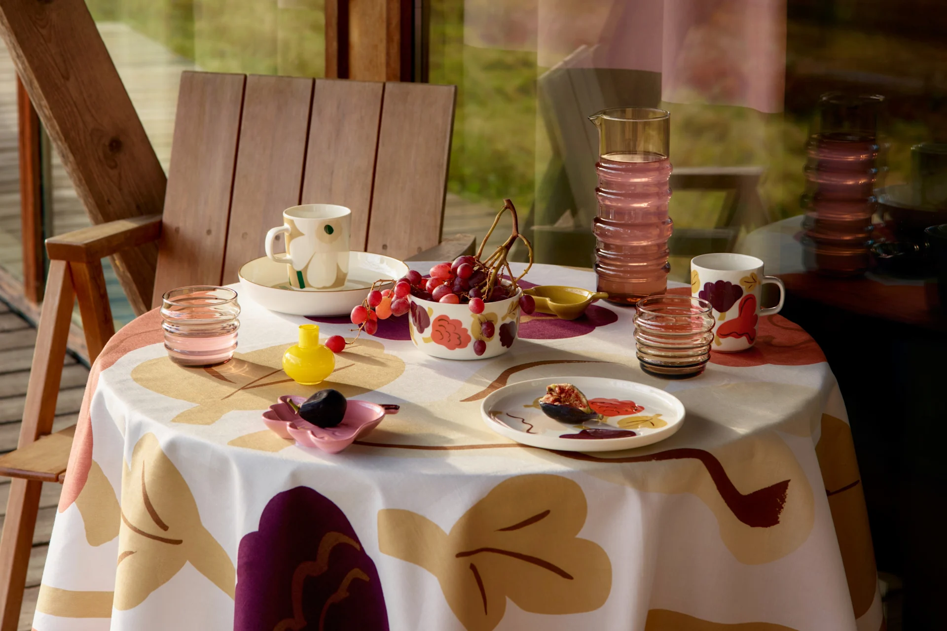A table set with a patterned tablecloth, mugs, bowls of grapes, a fig on a plate, a pitcher, and glasses, next to a wooden chair.