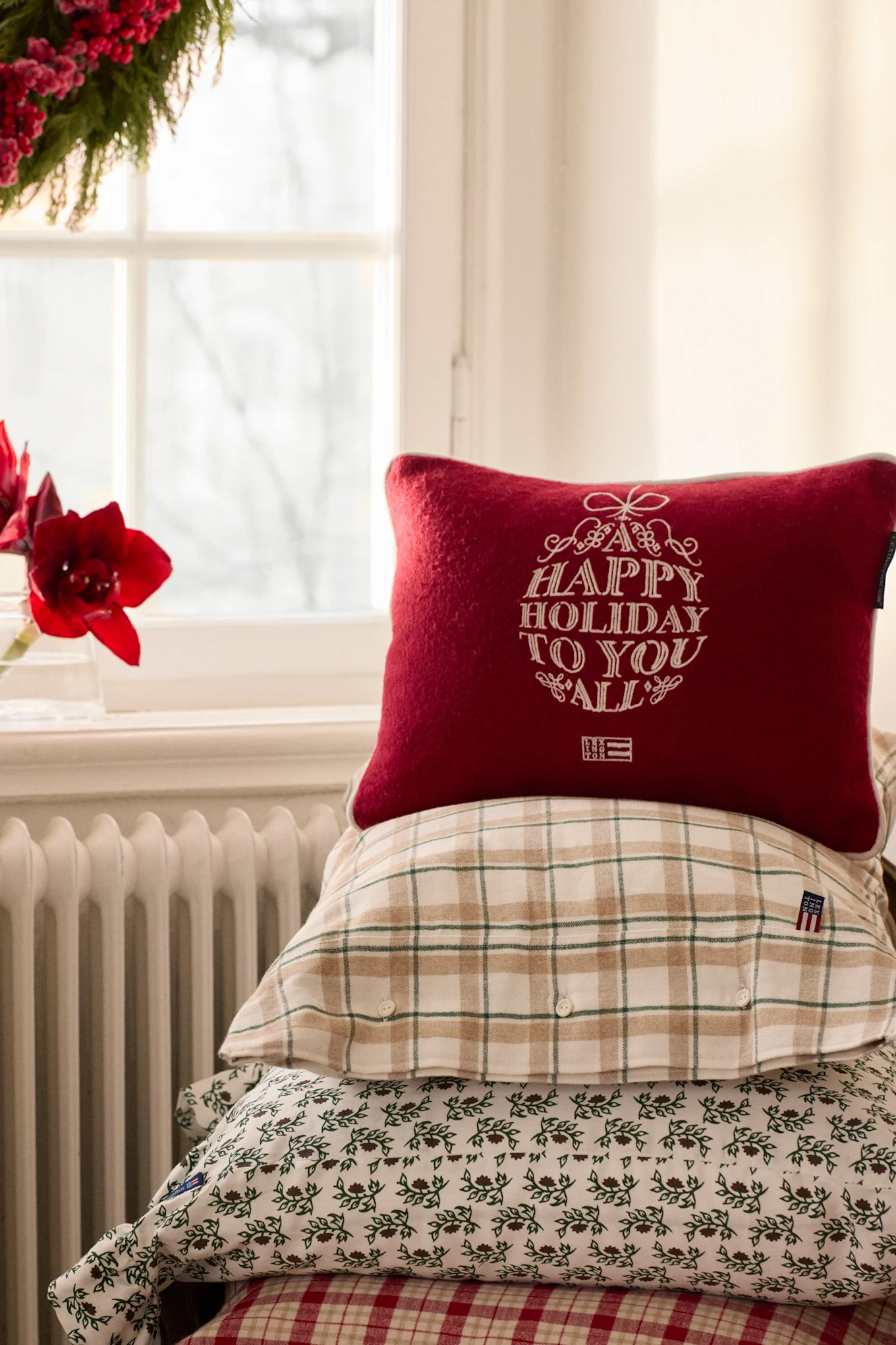 Three holiday pillows and a plaid blanket on a sofa, with a snow globe and Christmas tree lights nearby.