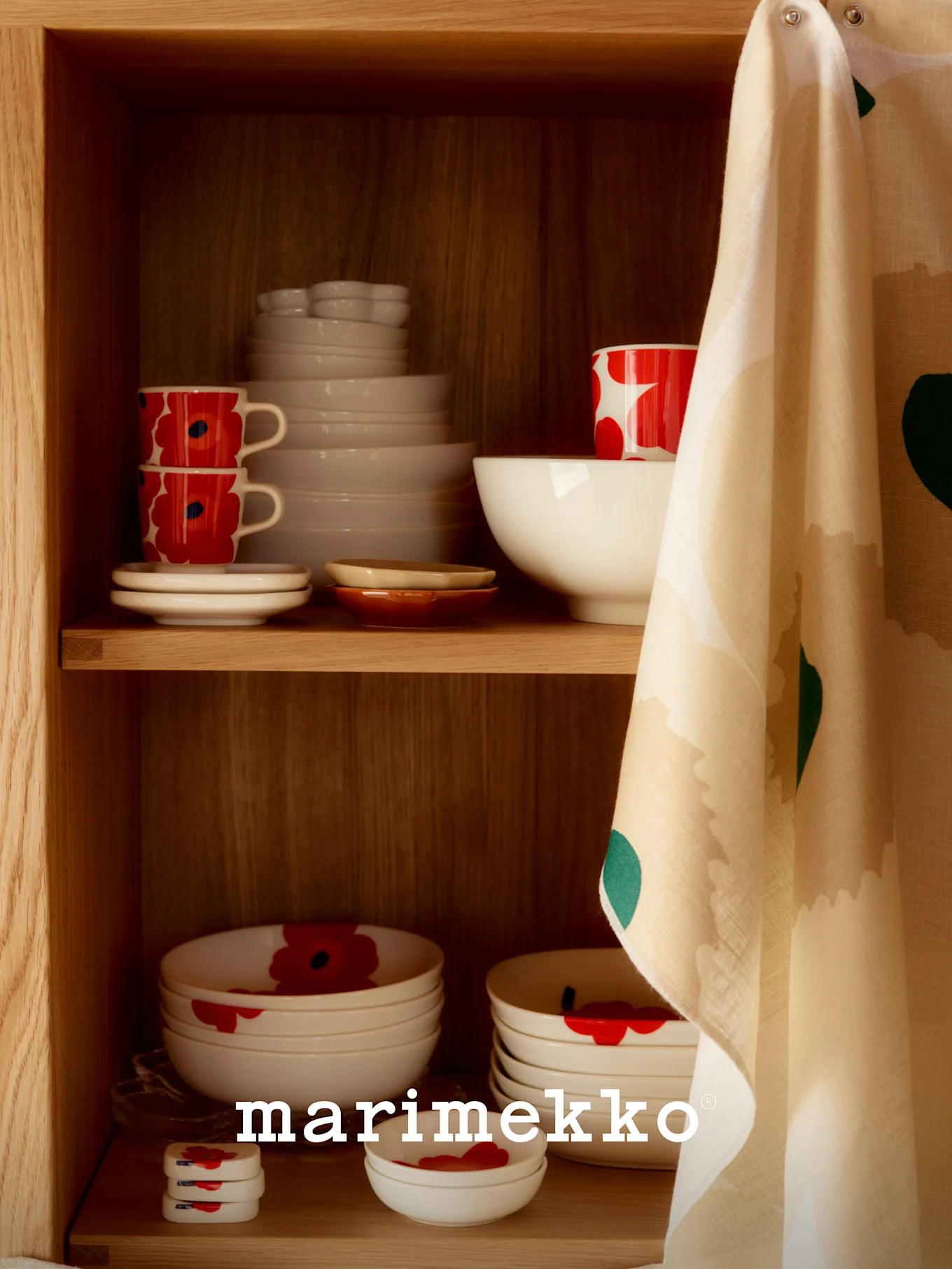 A wooden cabinet shelf with white ceramic dishes, some with a red poppy pattern, and a patterned tea towel hanging on the side.