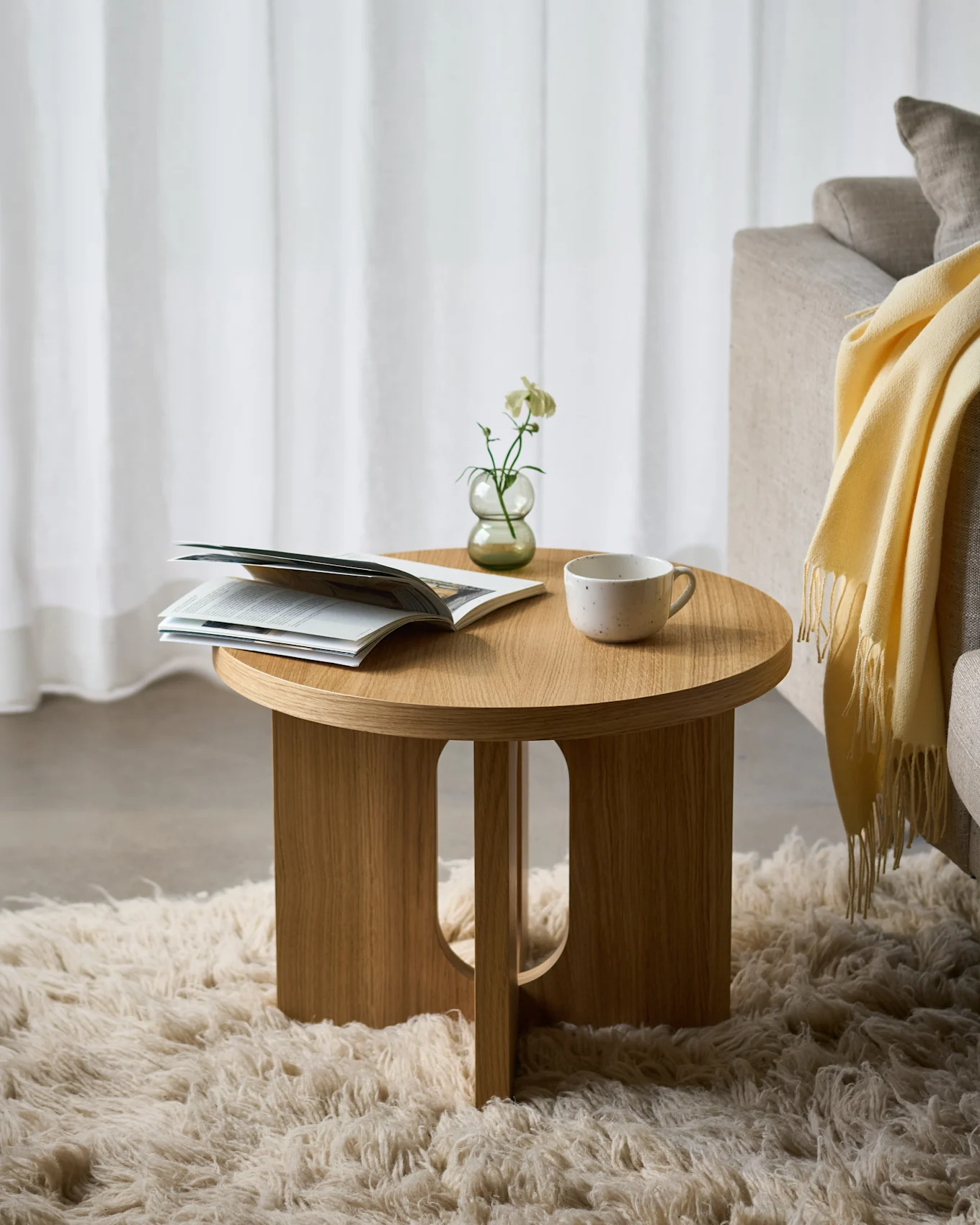 Wooden side table on a fluffy rug, holding an open magazine, a white flower in a vase, and a ceramic cup, next to a sofa.