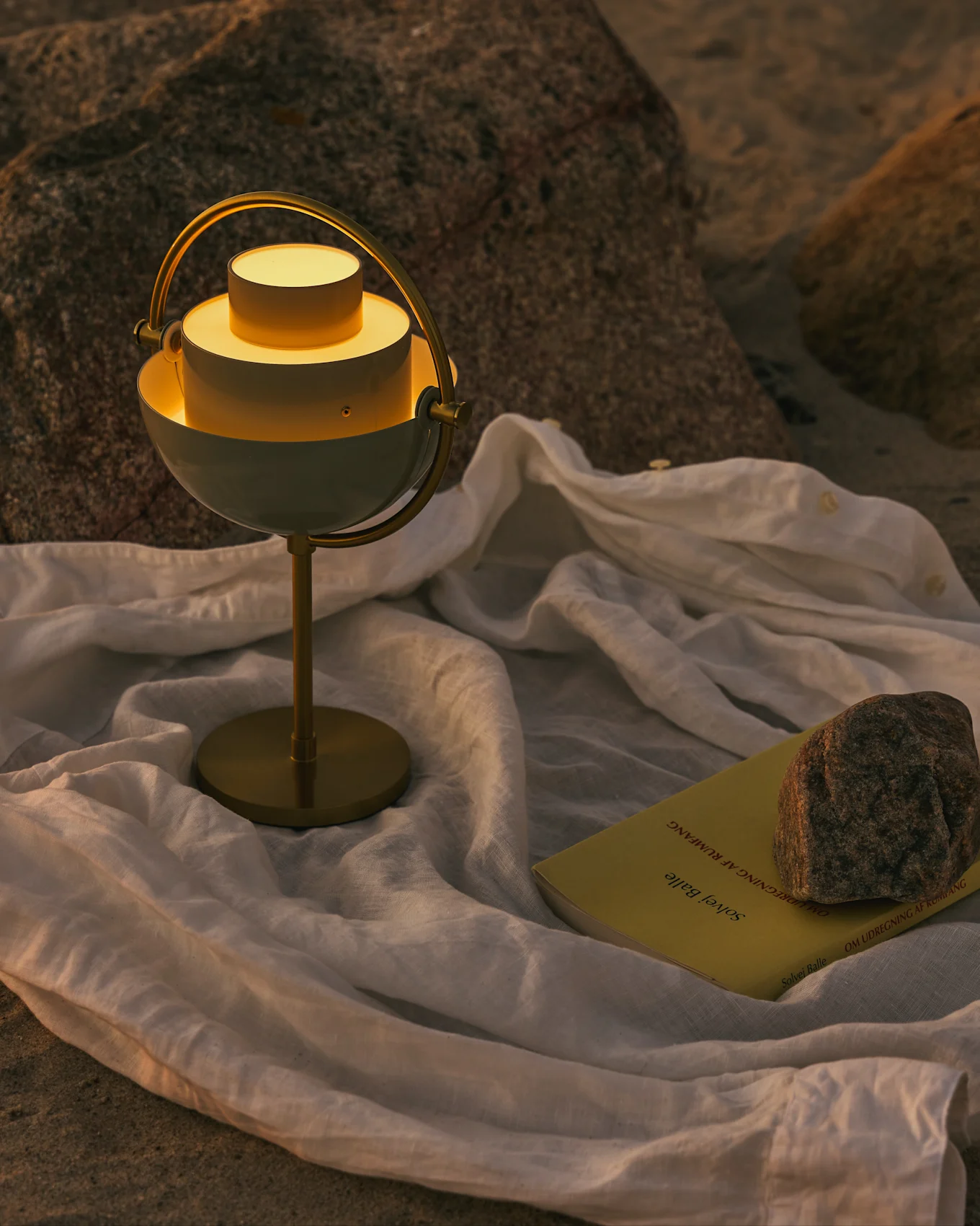 A modern white and brass table lamp with a glowing light, a yellow book, and a rock on a white cloth on a rocky surface.