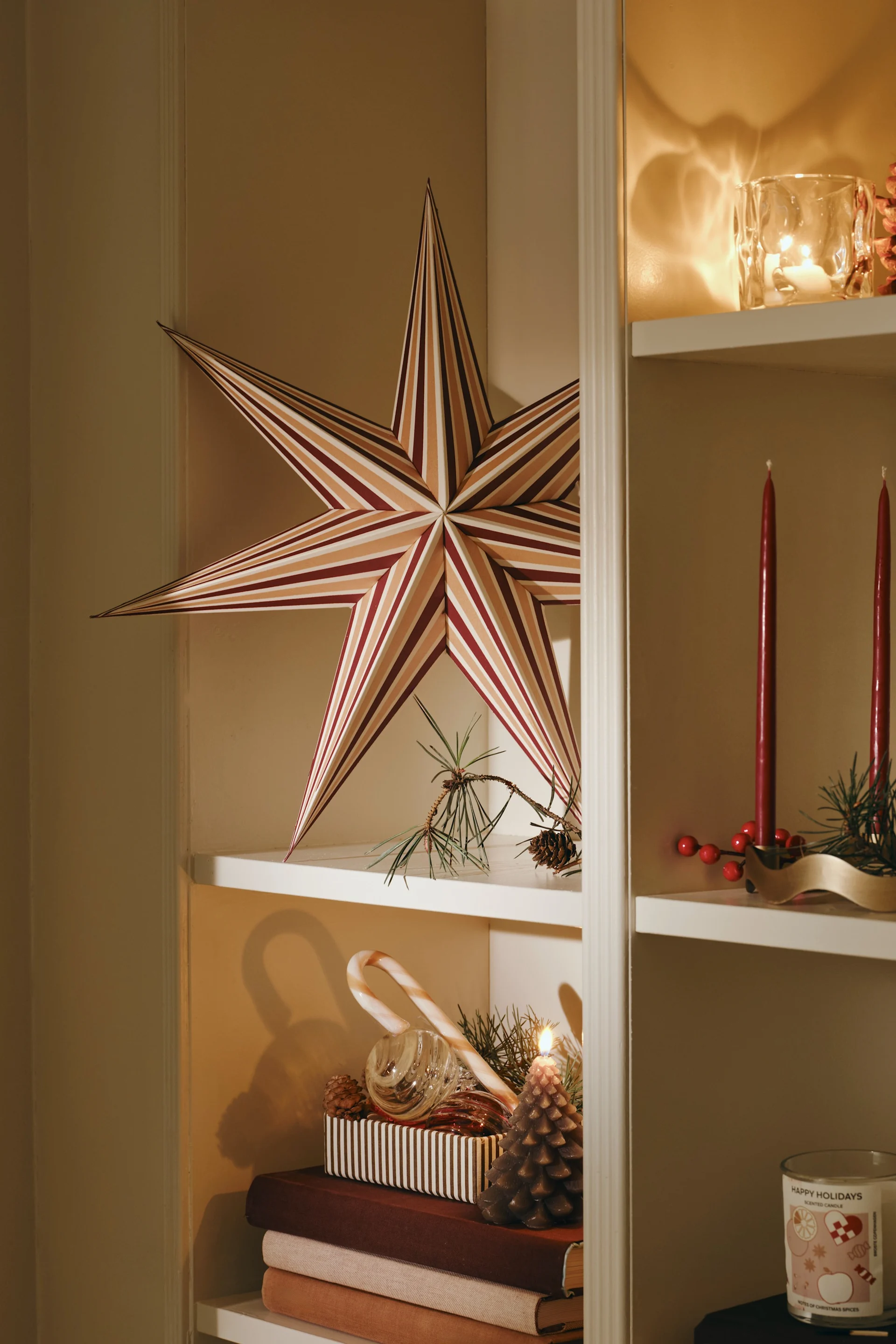 A white bookshelf displays holiday decor: a large striped paper star, lit candles, red berries, pinecones, and a stack of books.