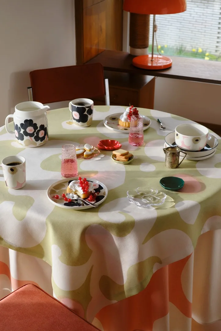A round table with a green, pink, and white floral tablecloth, set with black-and-pink patterned mugs, plates, a pitcher, pastries topped with red currants, and pink glasses.