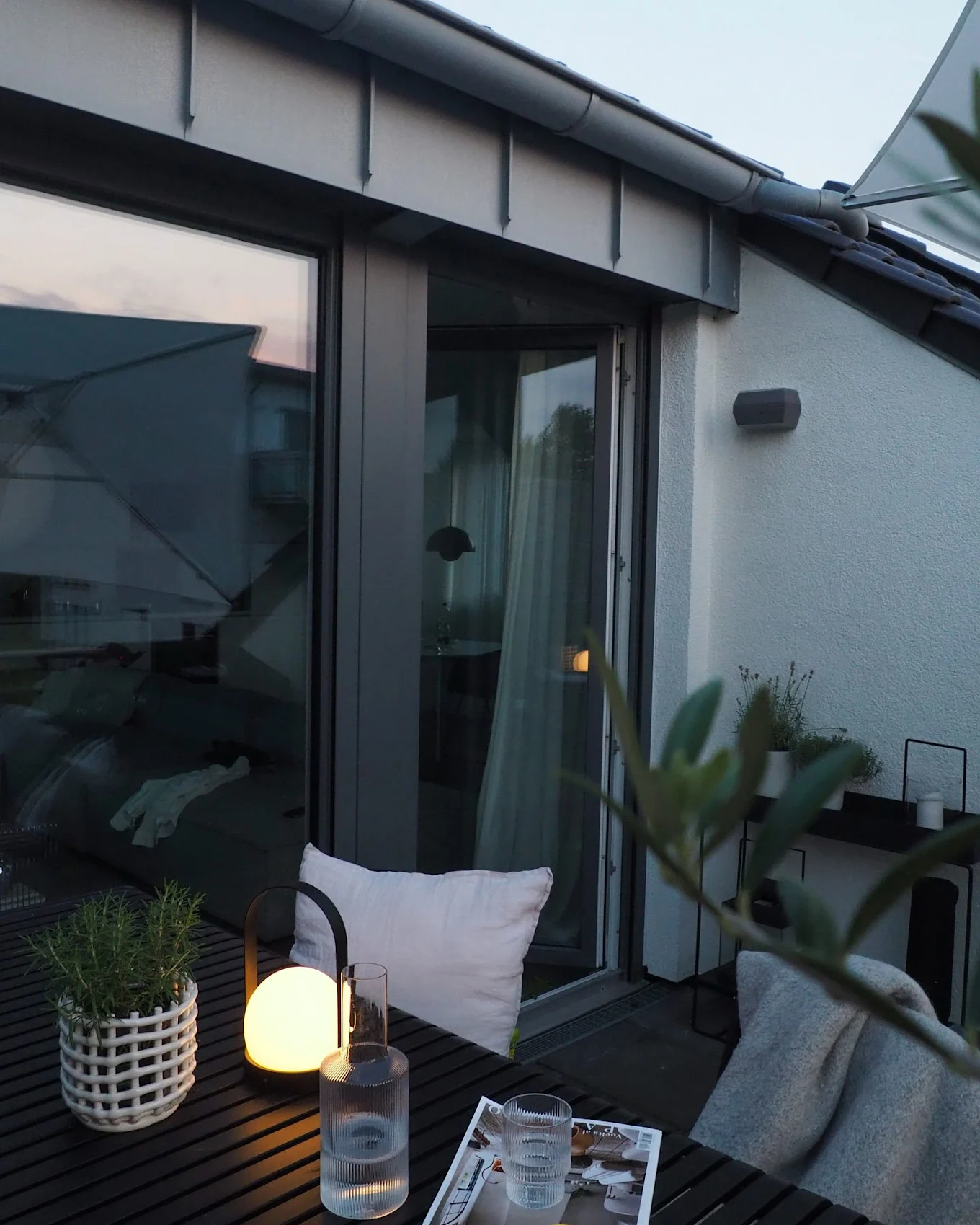 Evening balcony scene with a glowing lantern, a potted plant, and a glass of water on a dark slatted table.