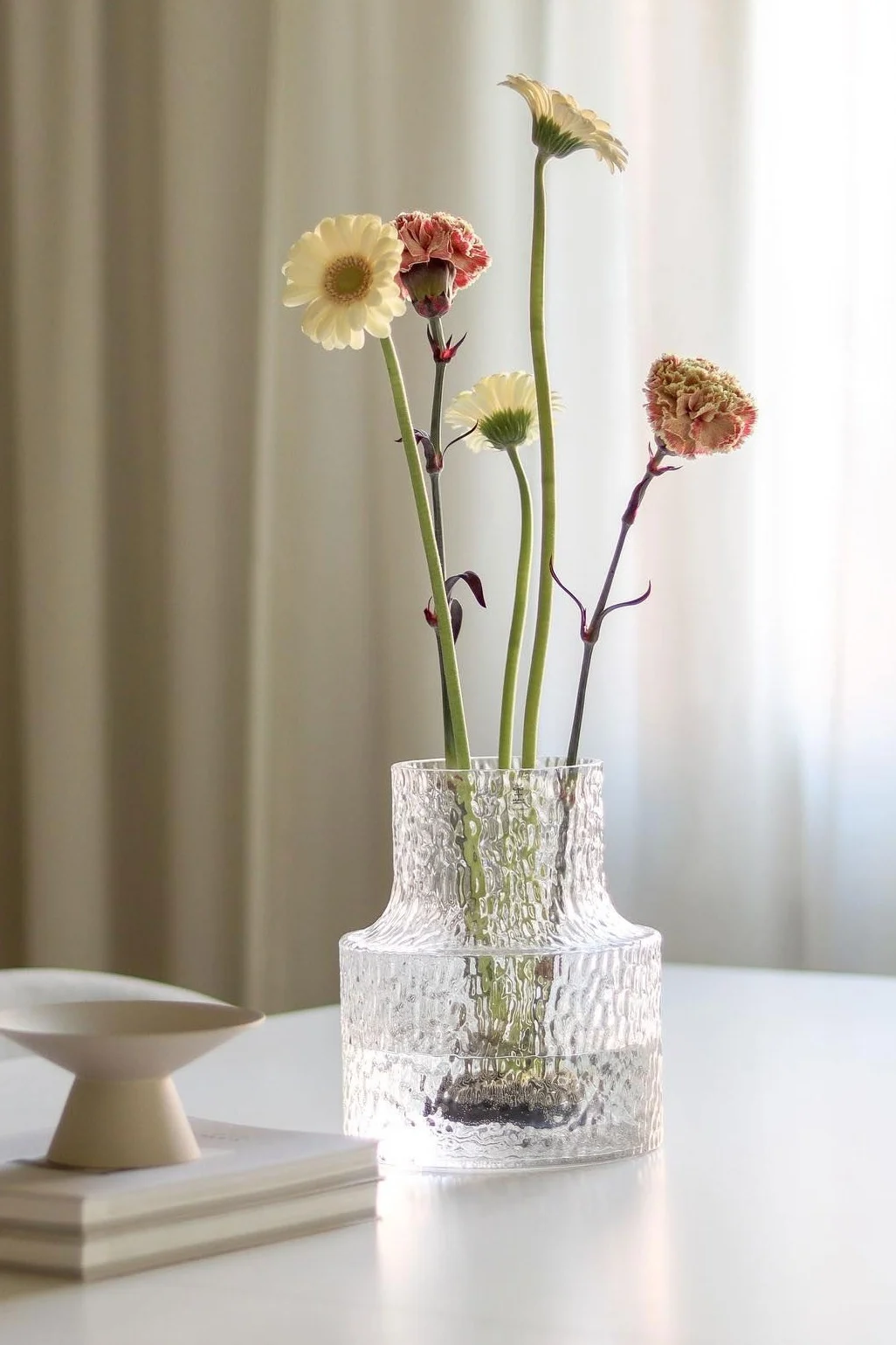 A clear, textured glass vase holds light yellow gerberas and reddish-pink carnations on a table.