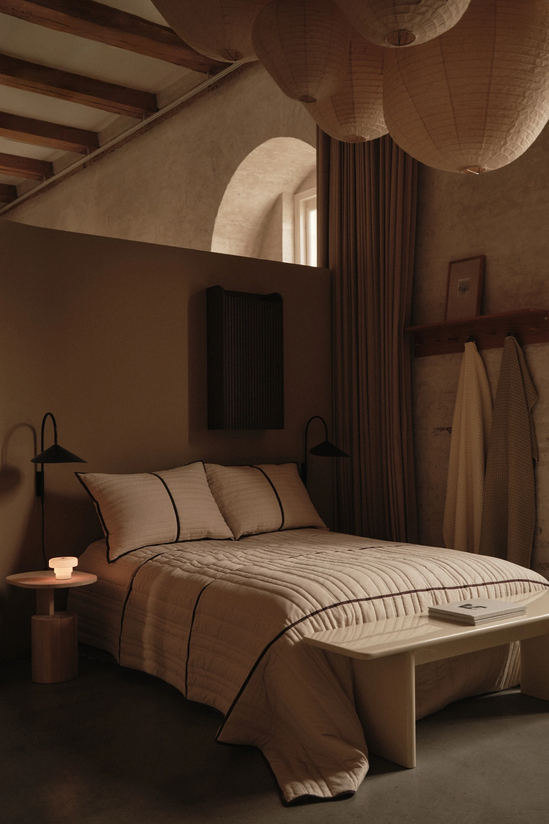 Cozy bedroom with a bed, side table, glowing lamp, and three large paper lanterns hanging from the ceiling.