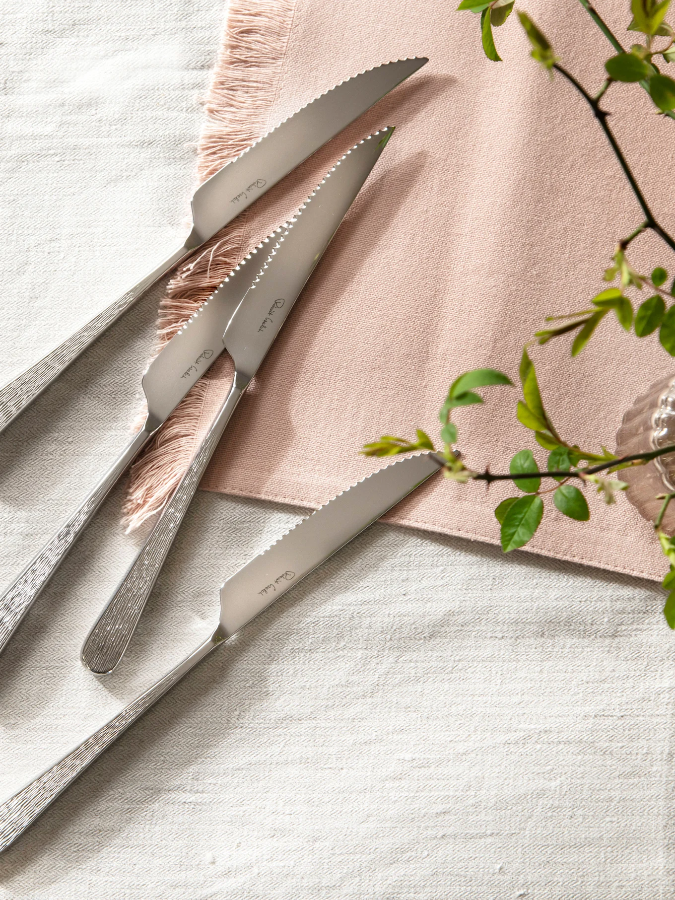 Four stainless steel steak knives with serrated blades and textured handles on a linen tablecloth with a pink napkin.