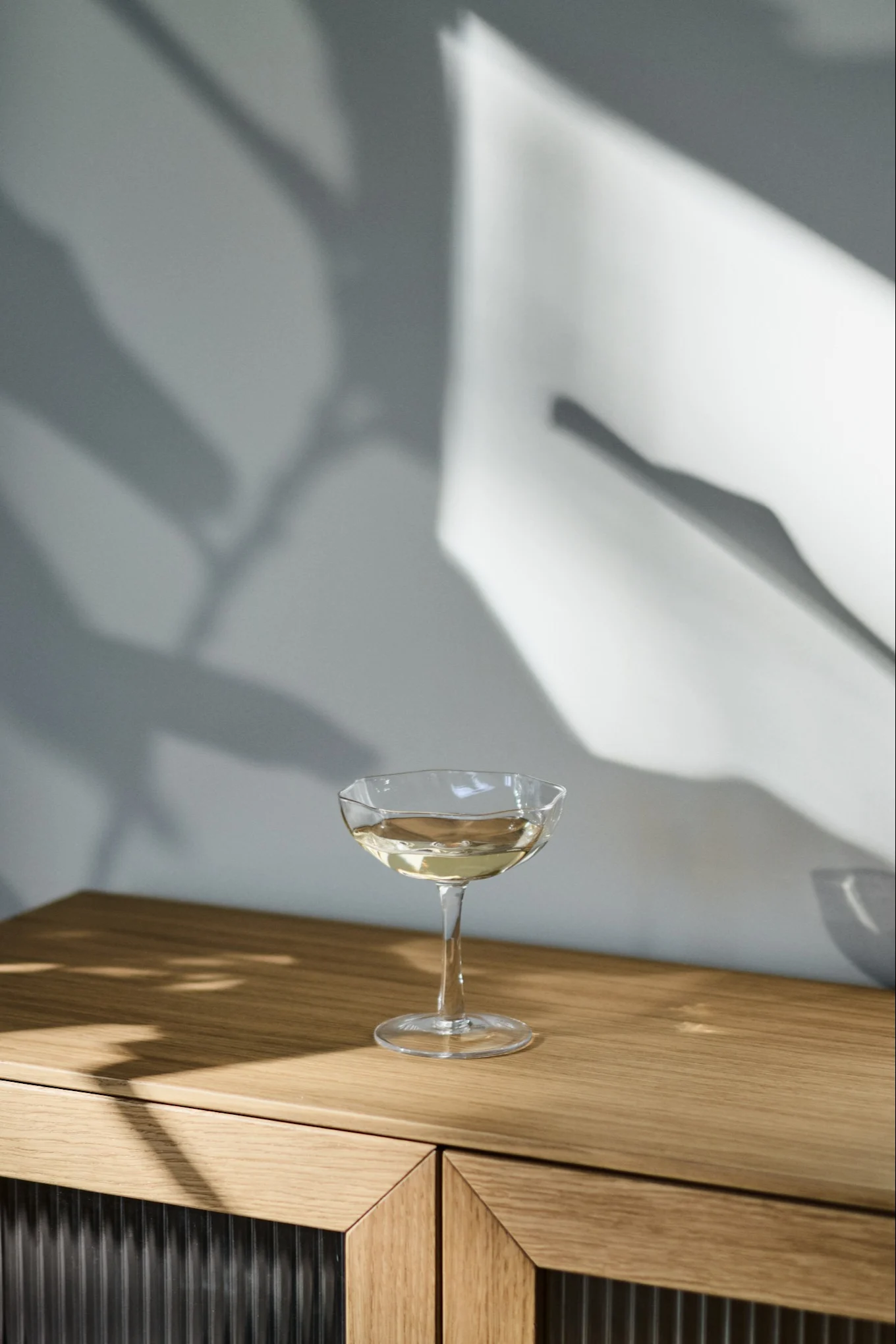 A coupe glass with a light drink on a wooden cabinet, with abstract shadows on the wall.