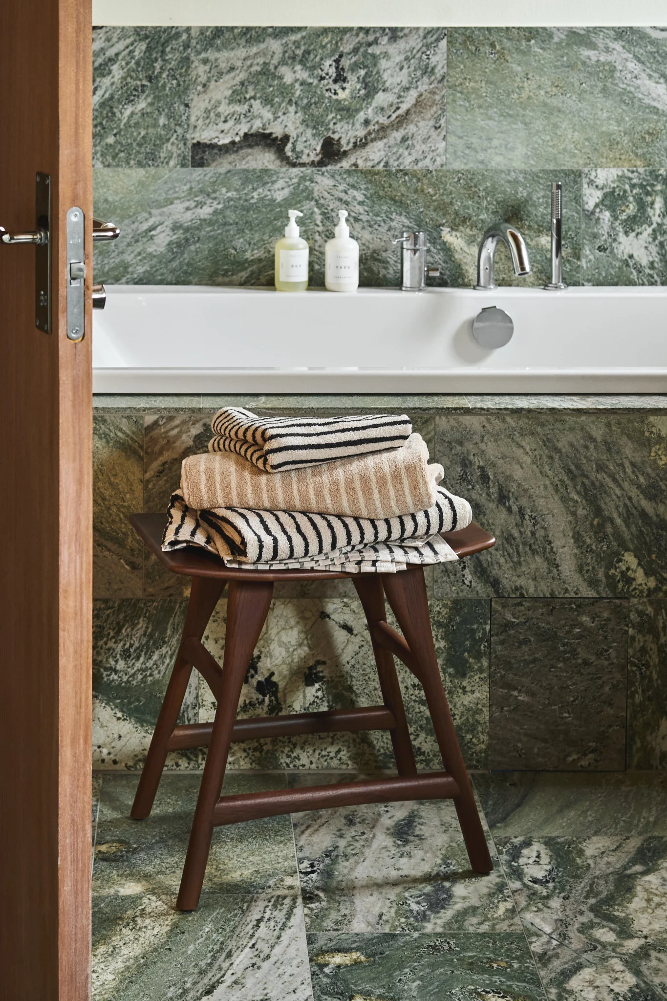 Bathroom with green marble tiles, white tub, and a wooden stool holding beige and off-white striped towels.