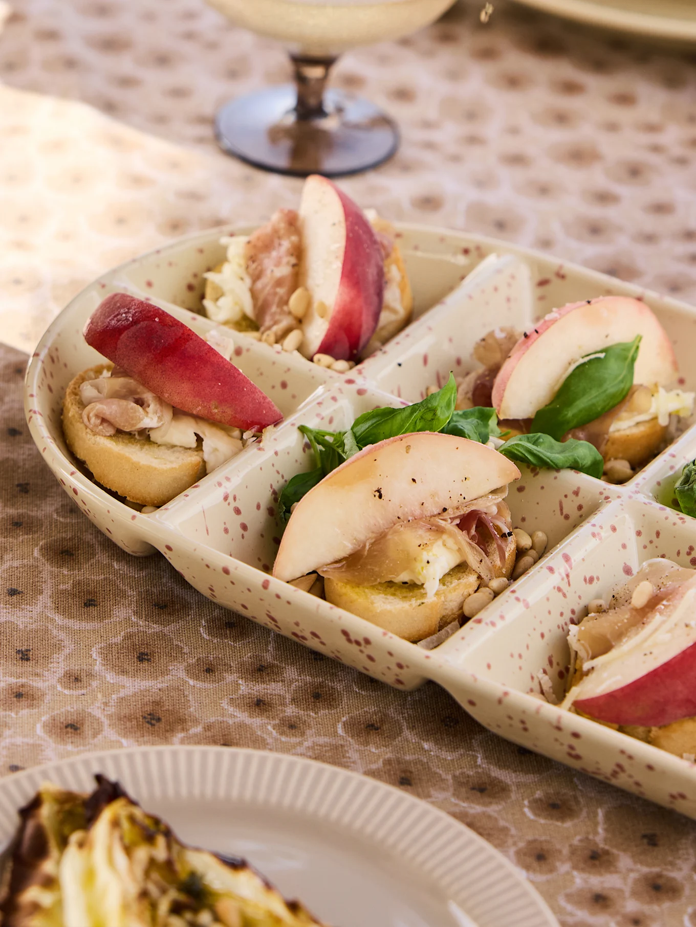 A segmented platter of appetizers with peach slices, prosciutto, basil, and pine nuts on small toasts.