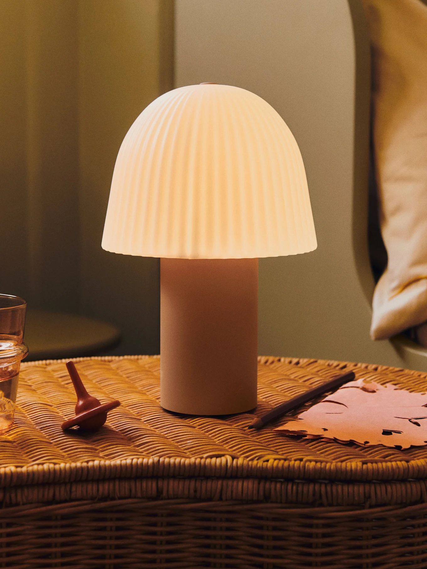 A glowing mushroom lamp with a pleated shade on a wicker bedside table with a glass of water, a spinning top, and paper.