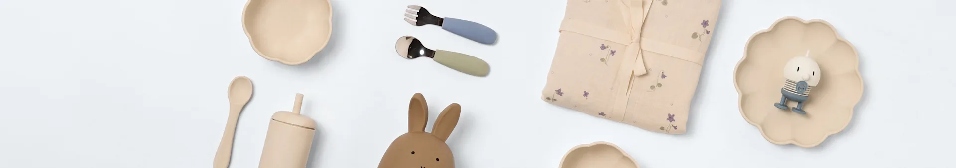 Flat lay of baby dining and play items: beige bowls, blue fork, green spoon, floral blanket, character candle on a plate, wooden spoon, bunny toy.