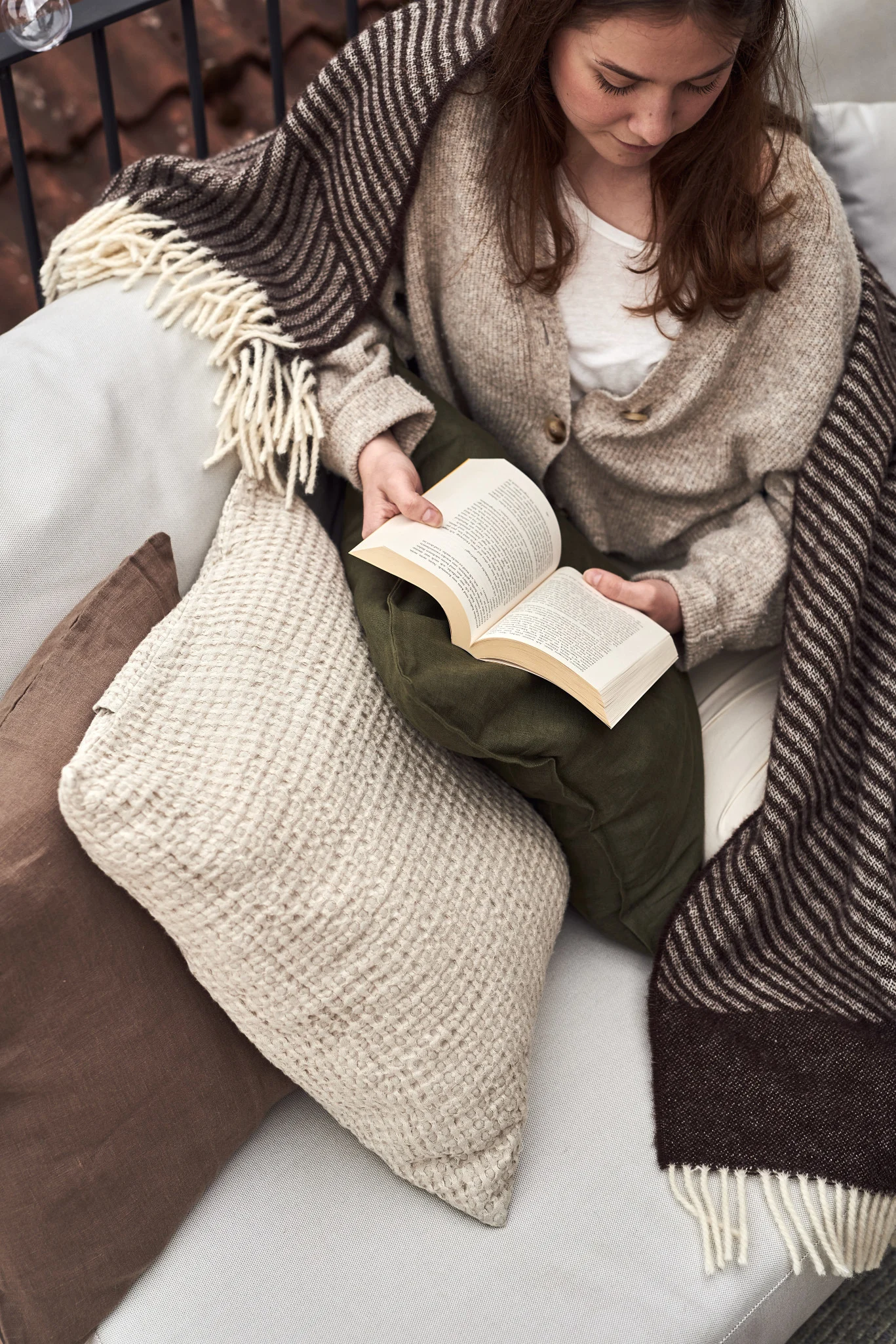 A woman is sitting on a sofa on the balcony reading a book.